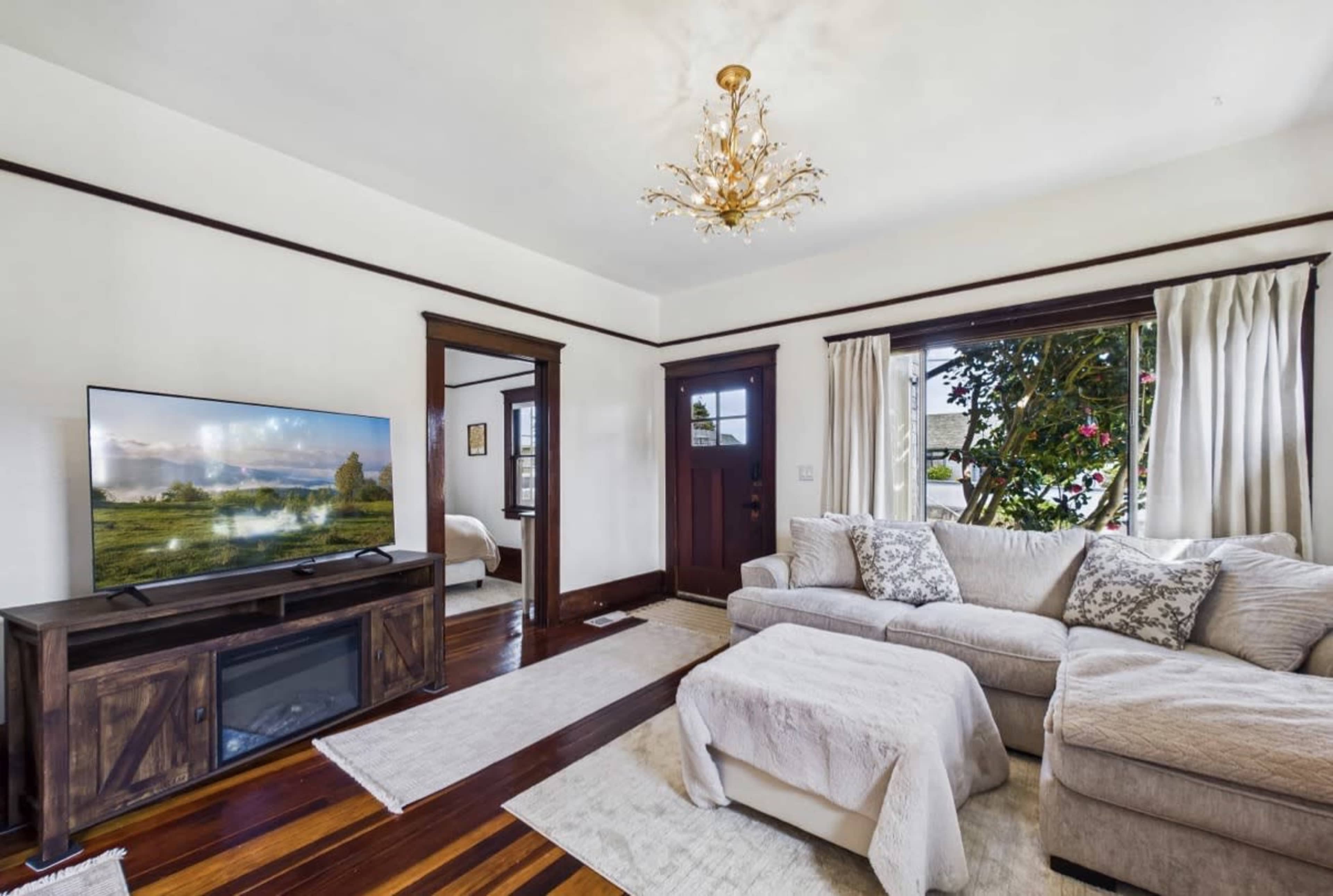 The living room features a sectional sofa, a television on a wooden stand, large windows with curtains, and hardwood floors.