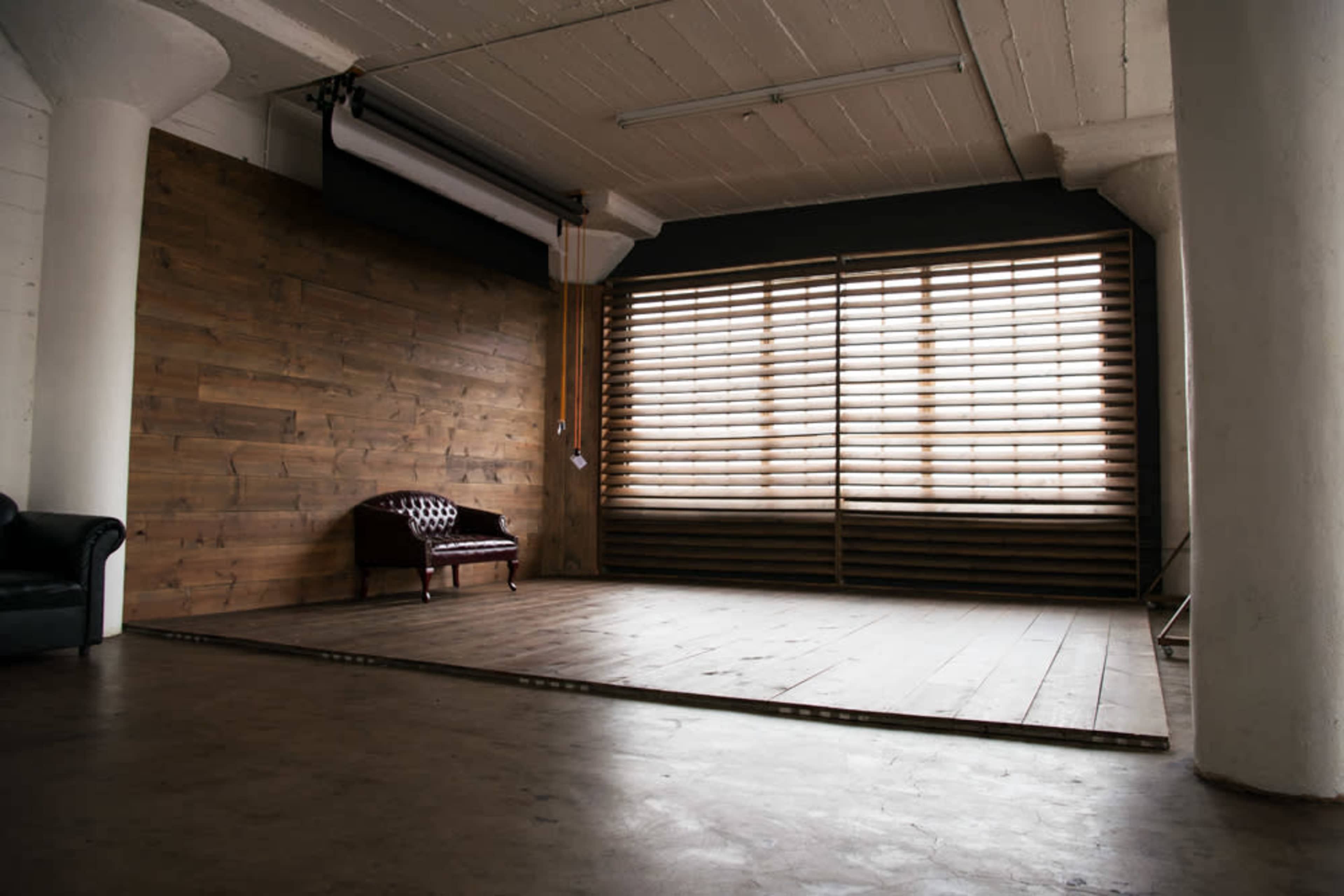 The image shows a large, empty studio space with wooden walls, a wooden floor, and a single leather couch near a window with horizontal blinds.