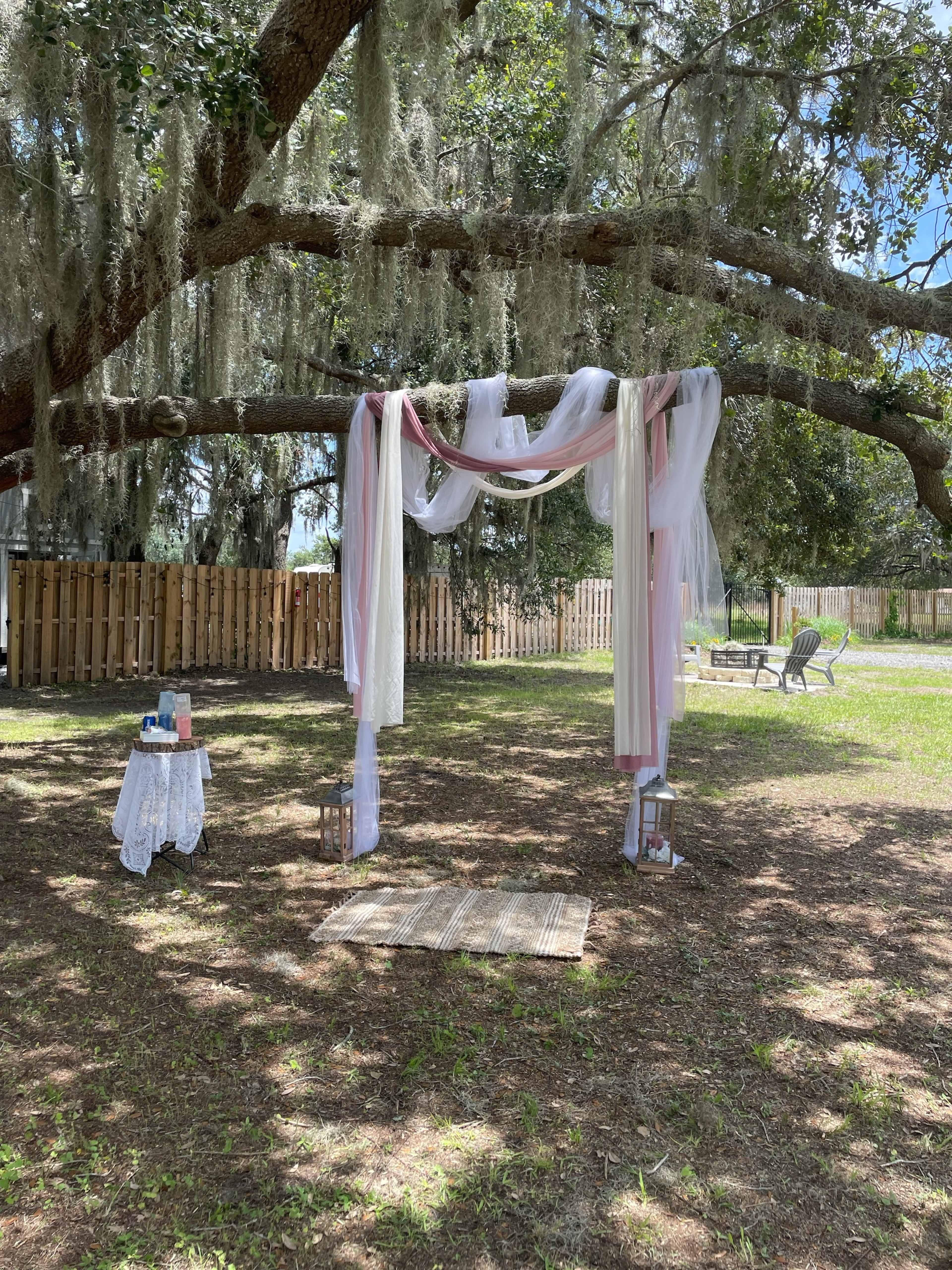 A decorated canopy with flowing fabric is set up under a tree, surrounded by lanterns and a table in a grassy area.