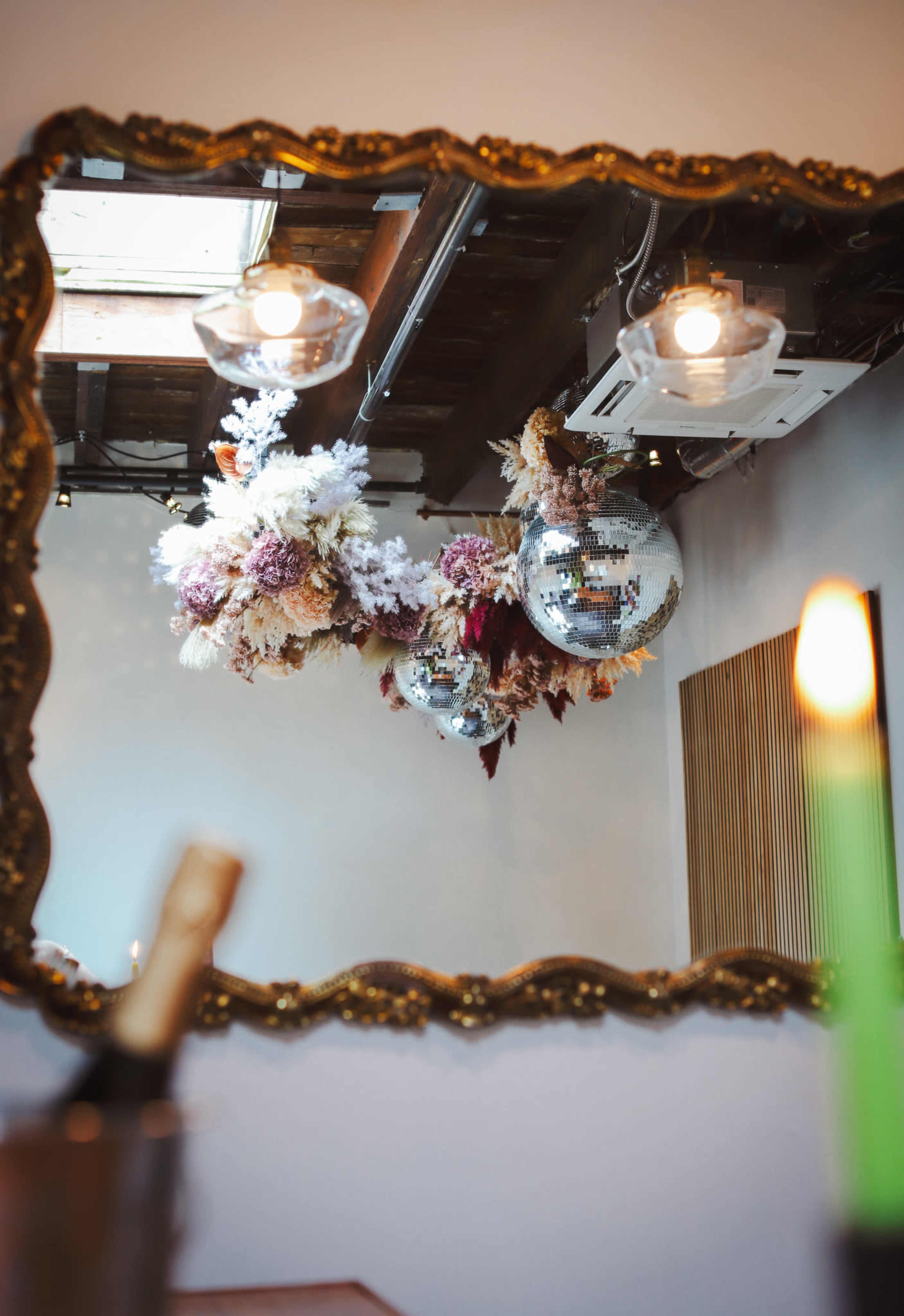 A decorative mirror reflects a ceiling adorned with floral arrangements and disco balls, with a candle in the foreground.