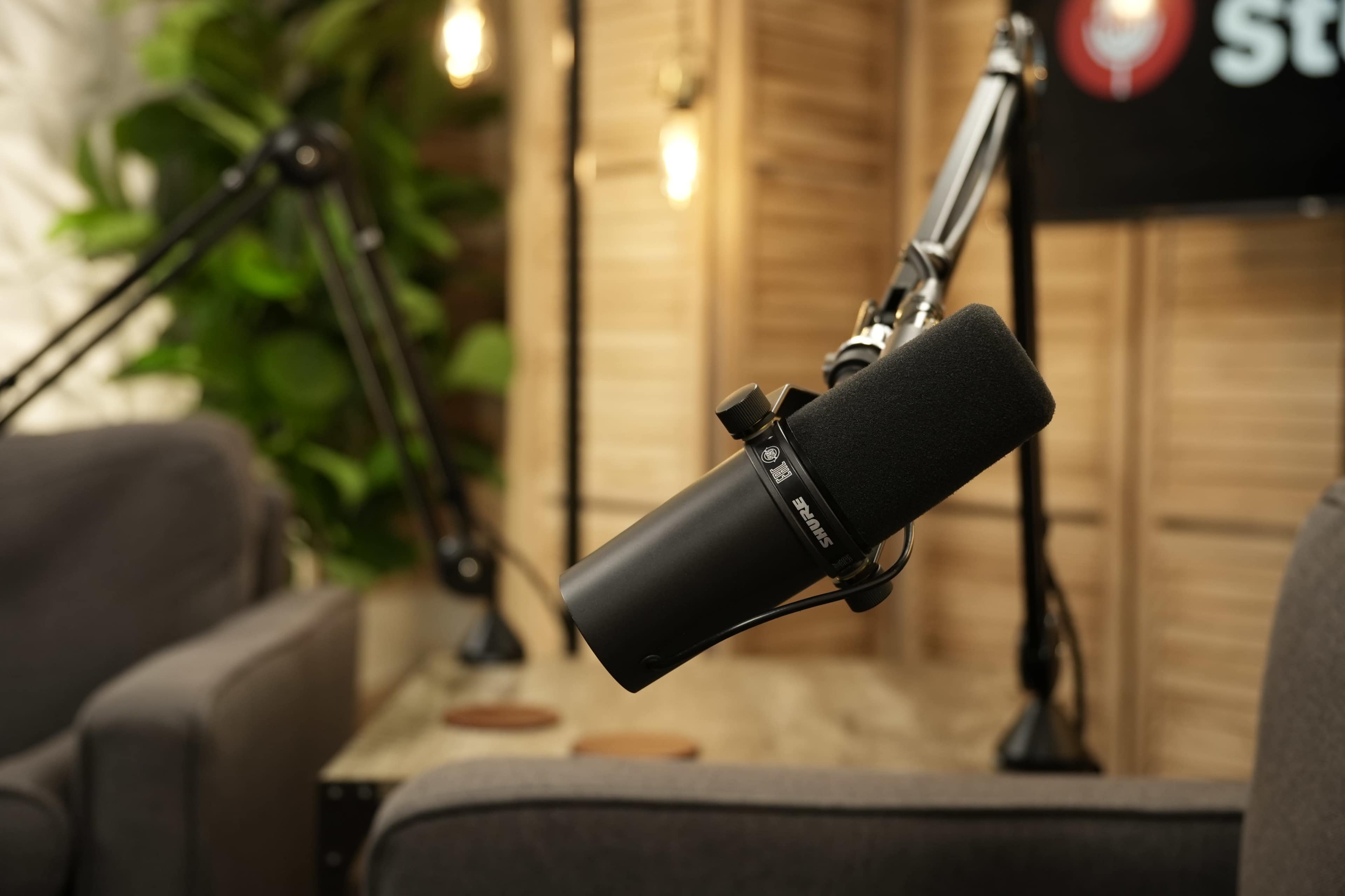 A black microphone is suspended on a boom arm above a gray couch in a well-lit recording studio.