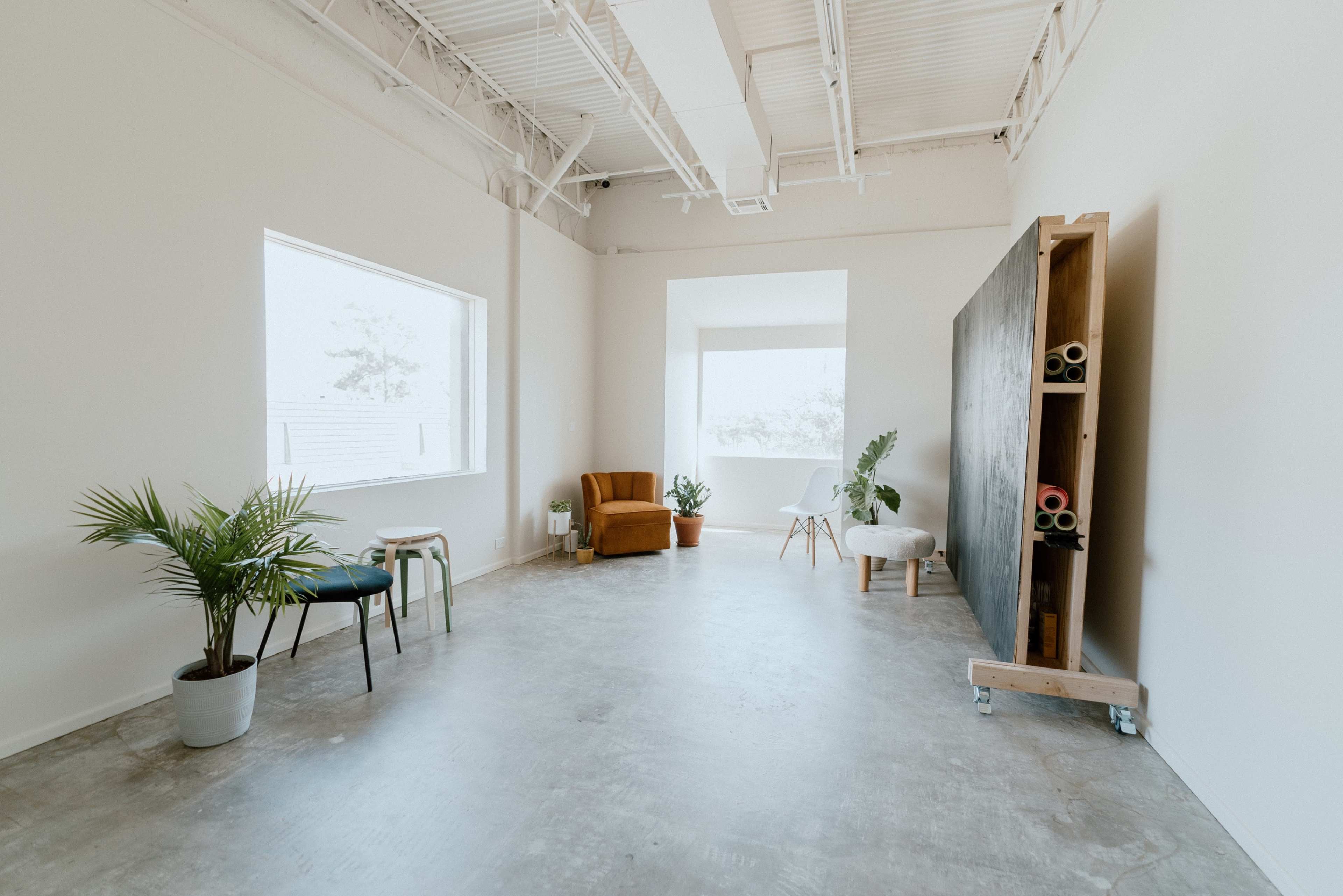 The image shows a spacious, minimalist room with a large window, a brown armchair, a small table, a plant, and a display of rolled materials against a wall.