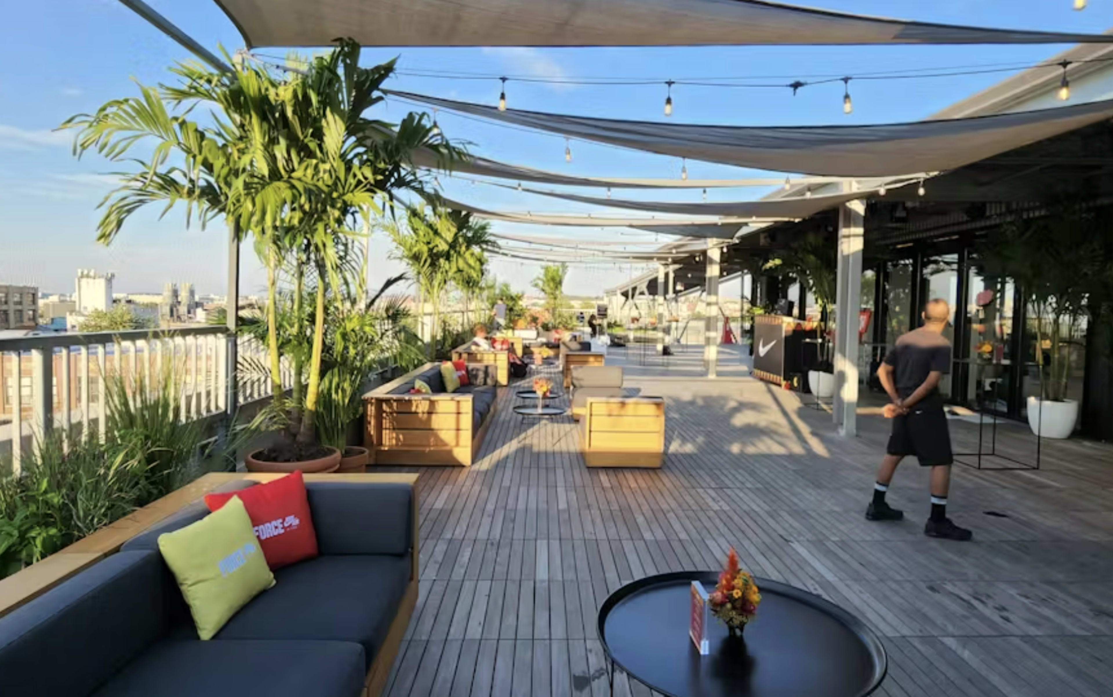 The image shows a spacious rooftop terrace with wooden flooring, seating areas, palm trees, and decorative string lights under a clear blue sky.