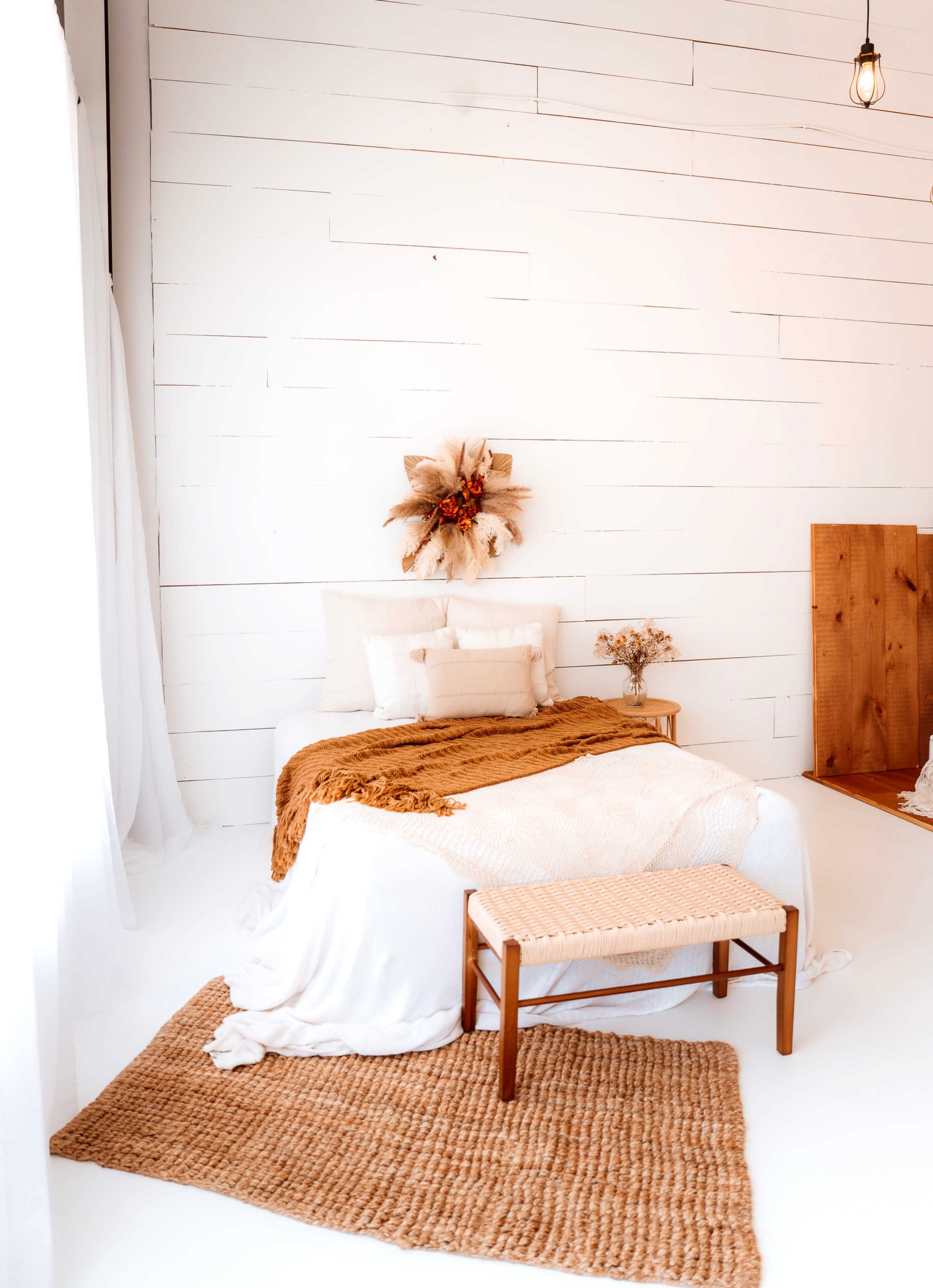 A neatly arranged bedroom features a white bed with textured blankets and pillows, a wooden bench at the foot, and decorative wall accents.