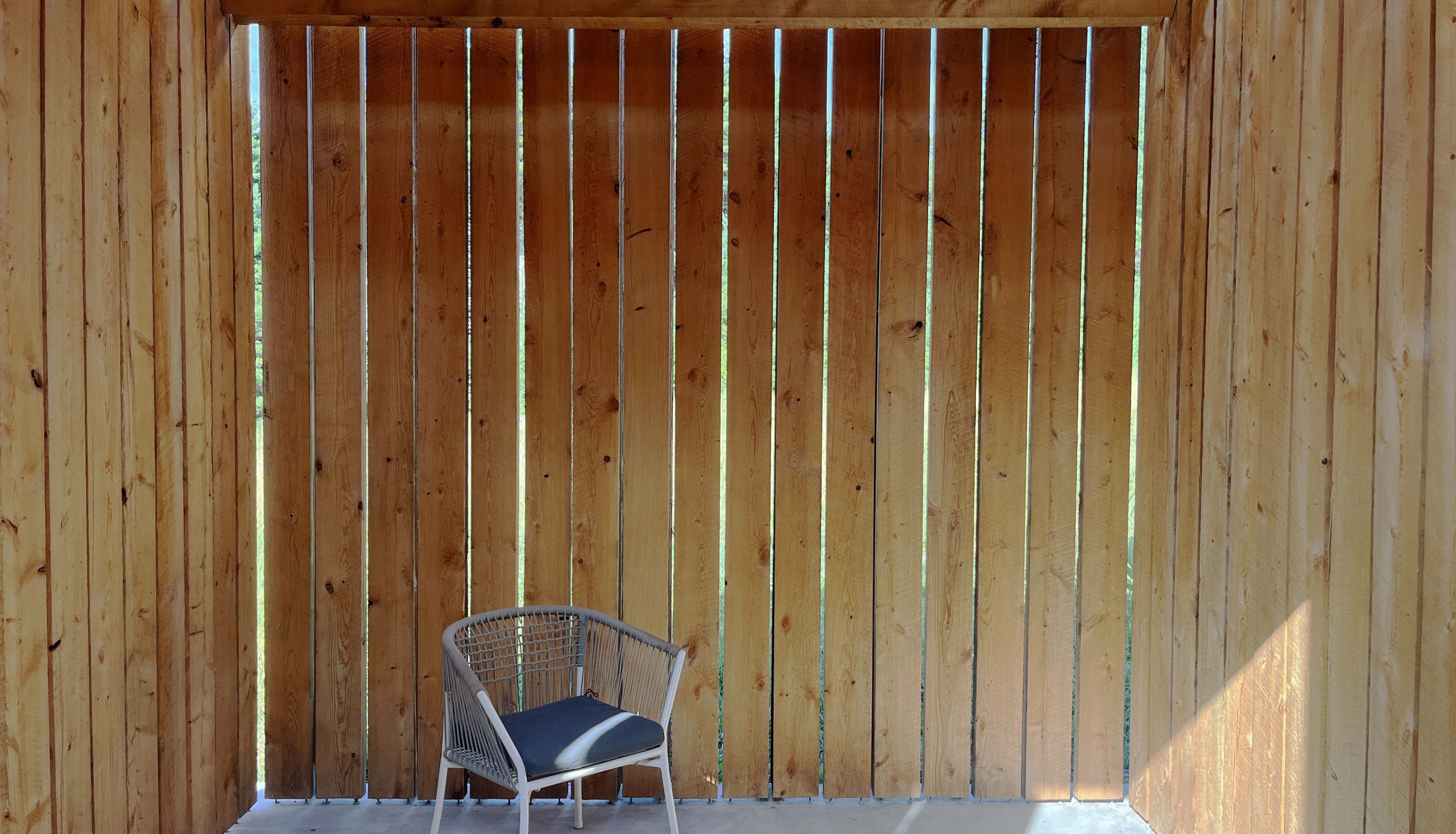 A single chair sits against a wooden wall with vertical slats, illuminated by natural light.