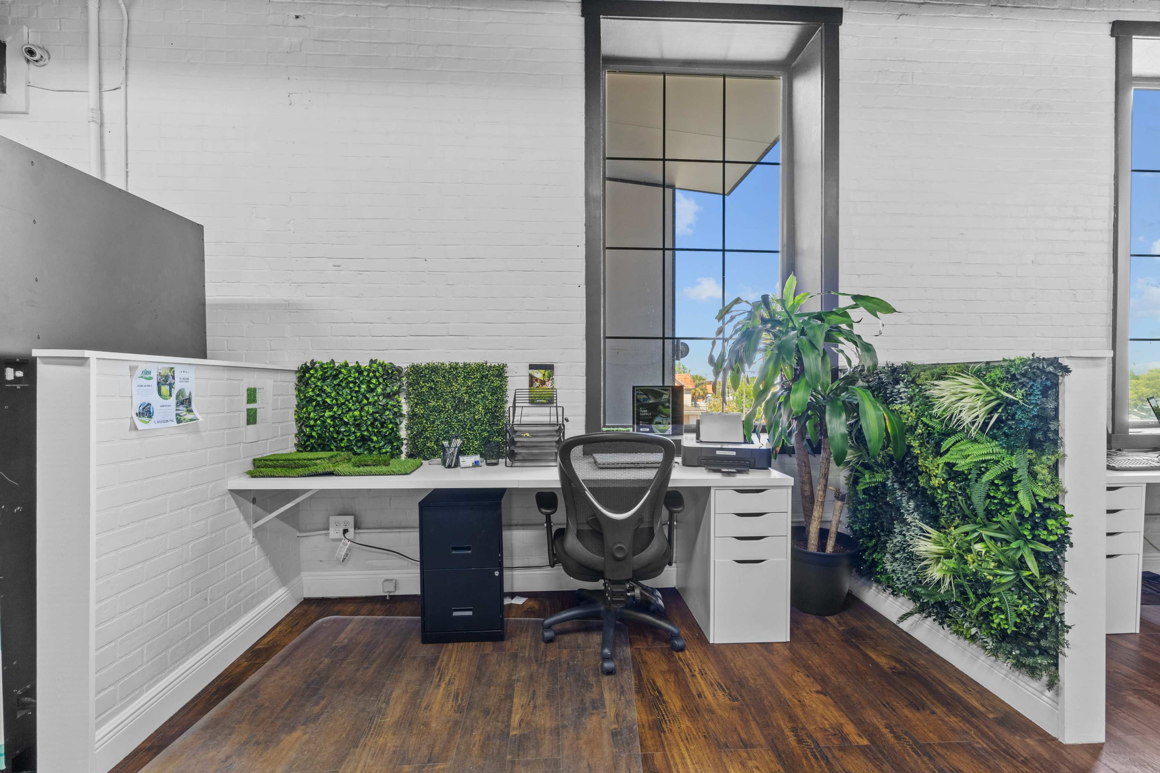 The image shows a modern office workspace featuring a black ergonomic chair, a white desk with drawers, and greenery on the walls beside a large window that lets in natural light.