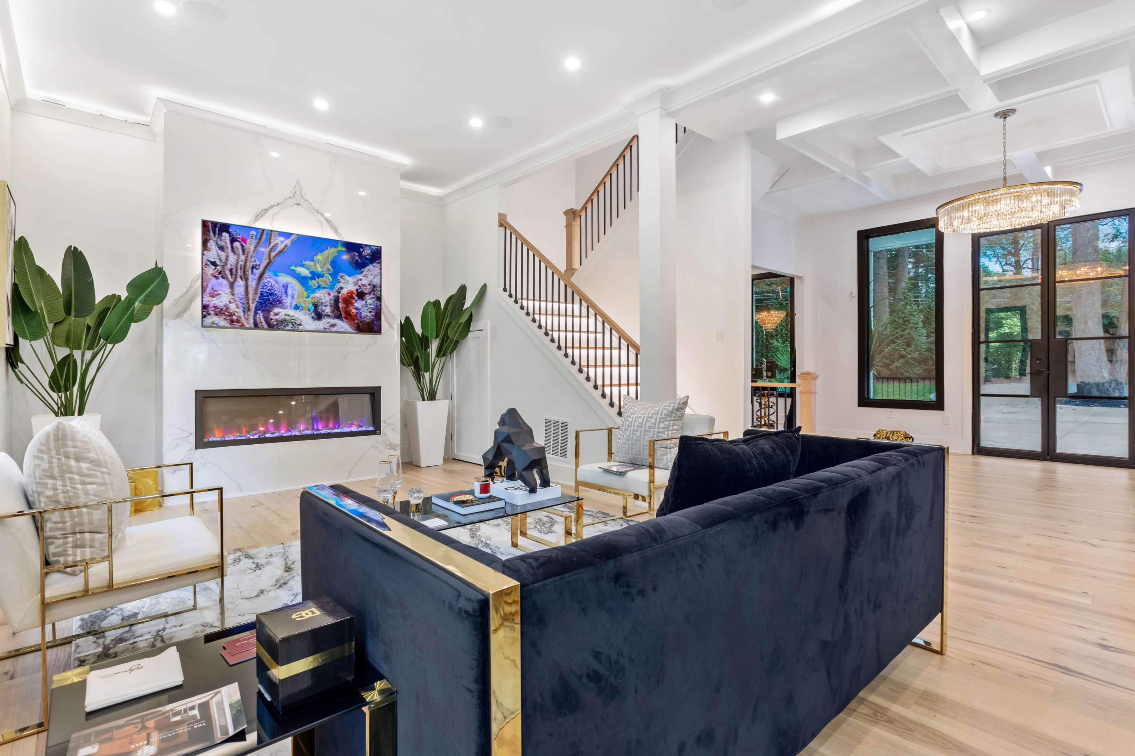 A modern living room features a navy blue sofa, a large flat-screen TV displaying an aquarium, a sleek fireplace, and a staircase leading to an upper level.