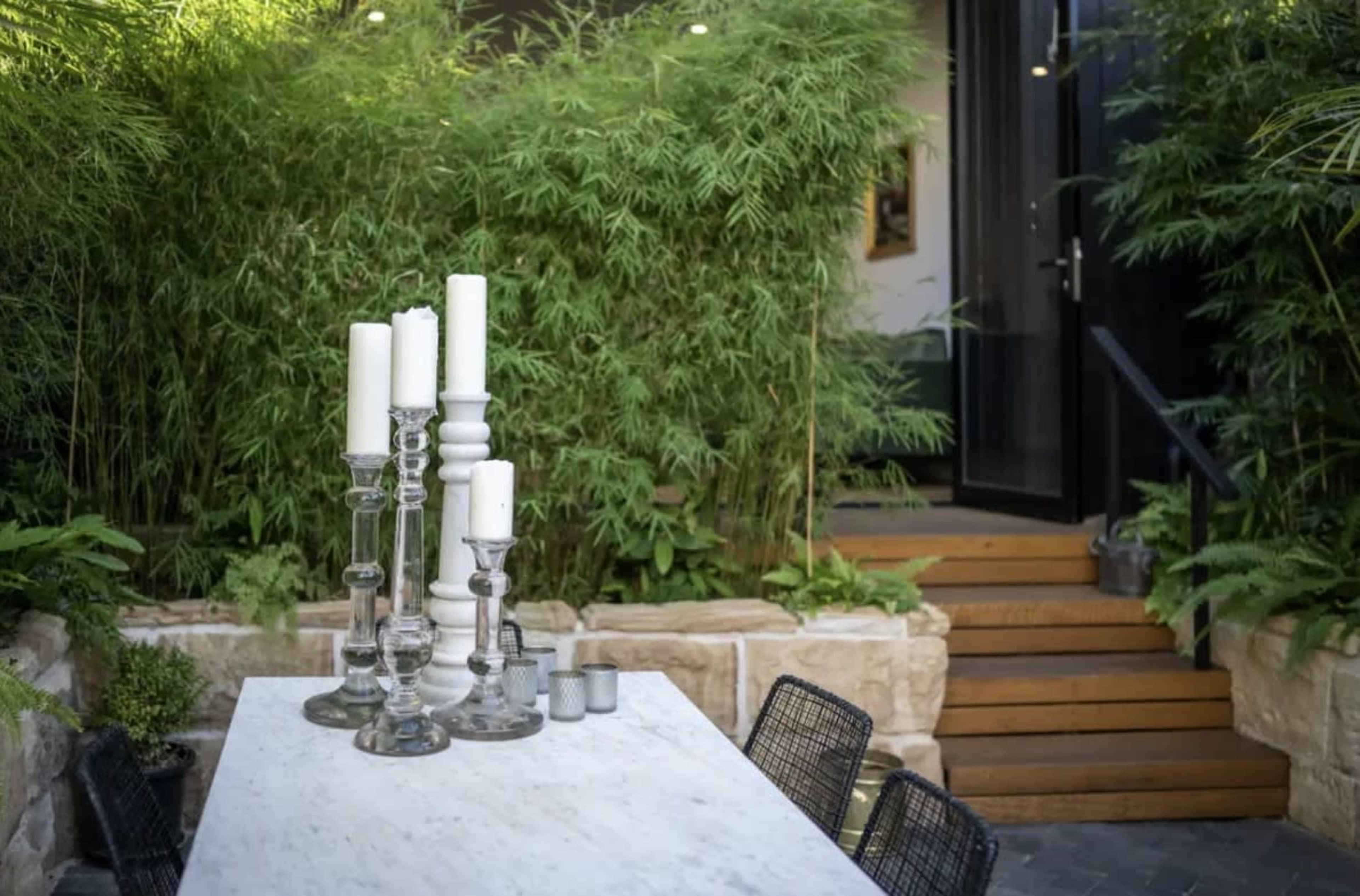 A stone table with tall candle holders stands in a lush green garden, adjacent to a set of wooden steps leading to a doorway.