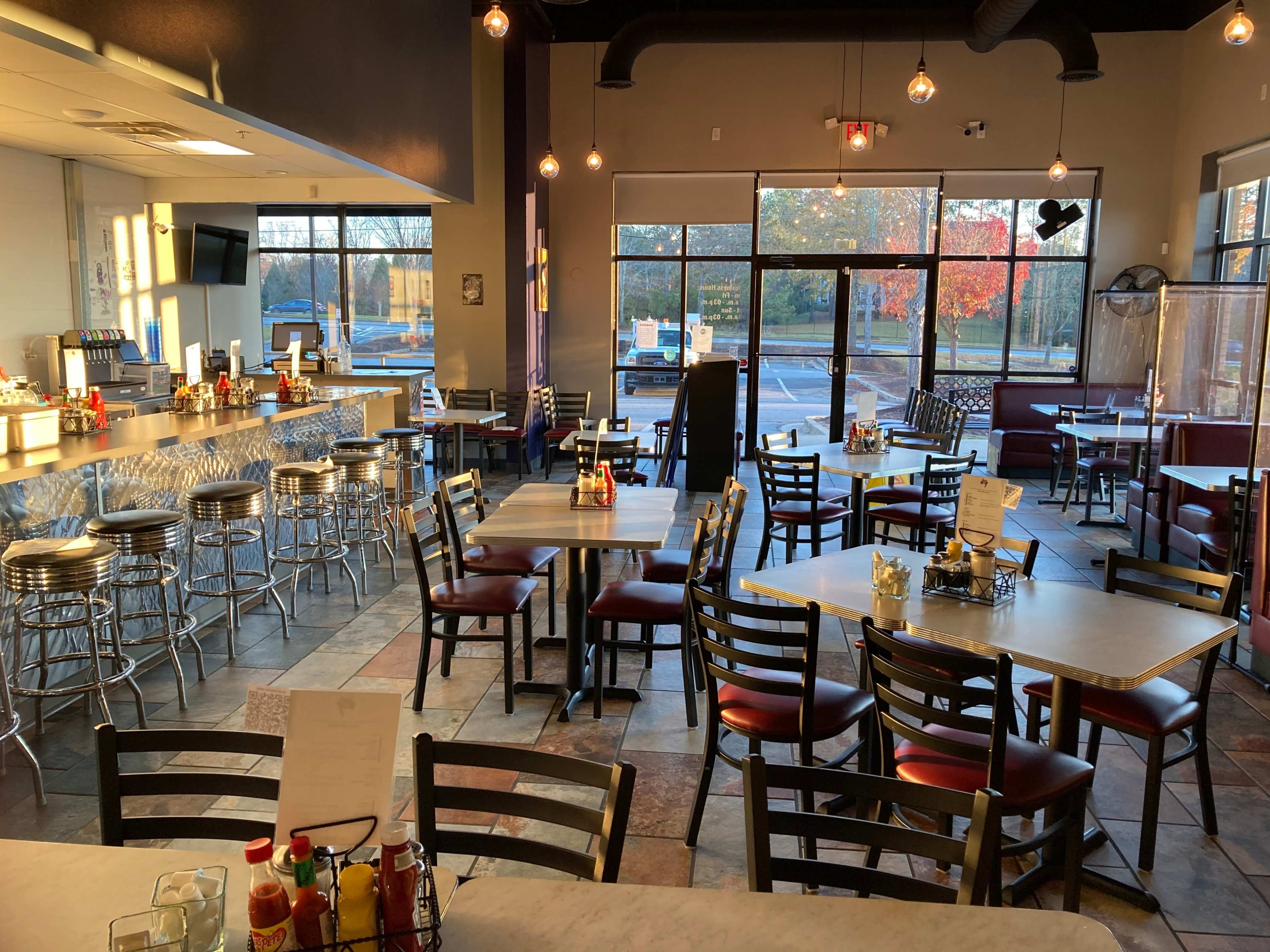 The image shows a modern restaurant interior with empty tables and chairs, a counter area, and large windows displaying an outdoor view.