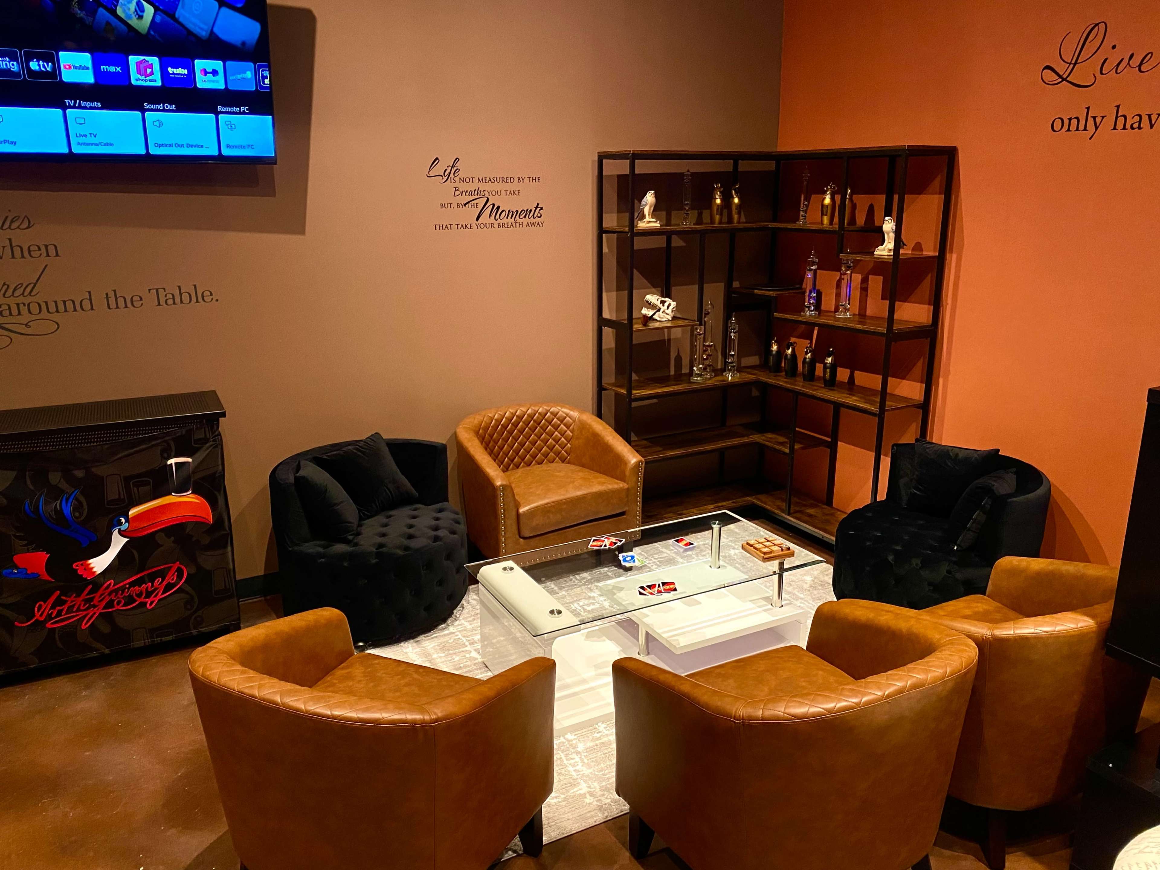 The image shows a cozy seating area with four leather chairs surrounding a central glass table, next to a wooden shelf displaying various decorative items.
