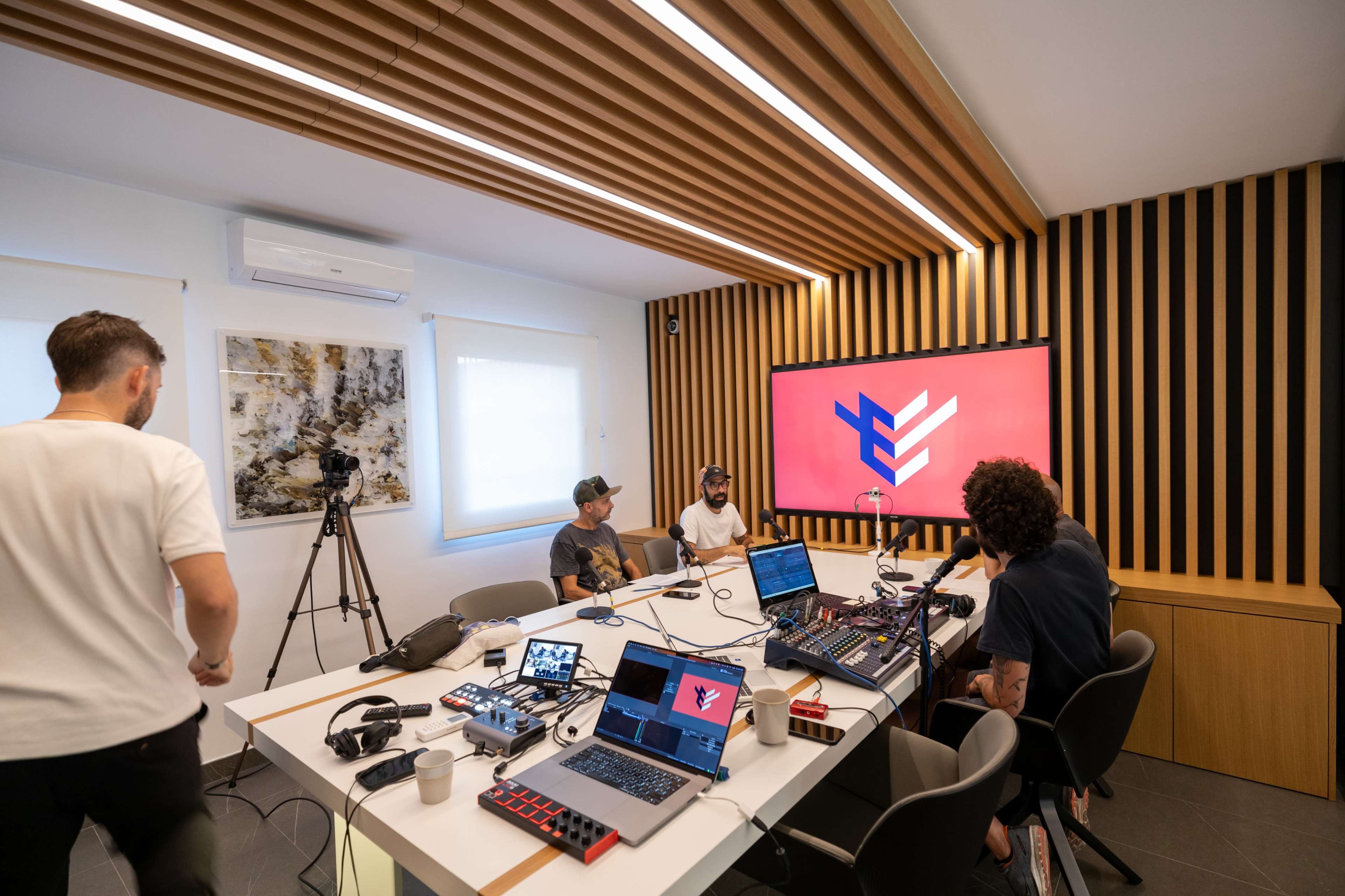 Four individuals are seated around a table in a modern studio equipped with audio and video equipment, while a large screen displays a graphic with a logo.