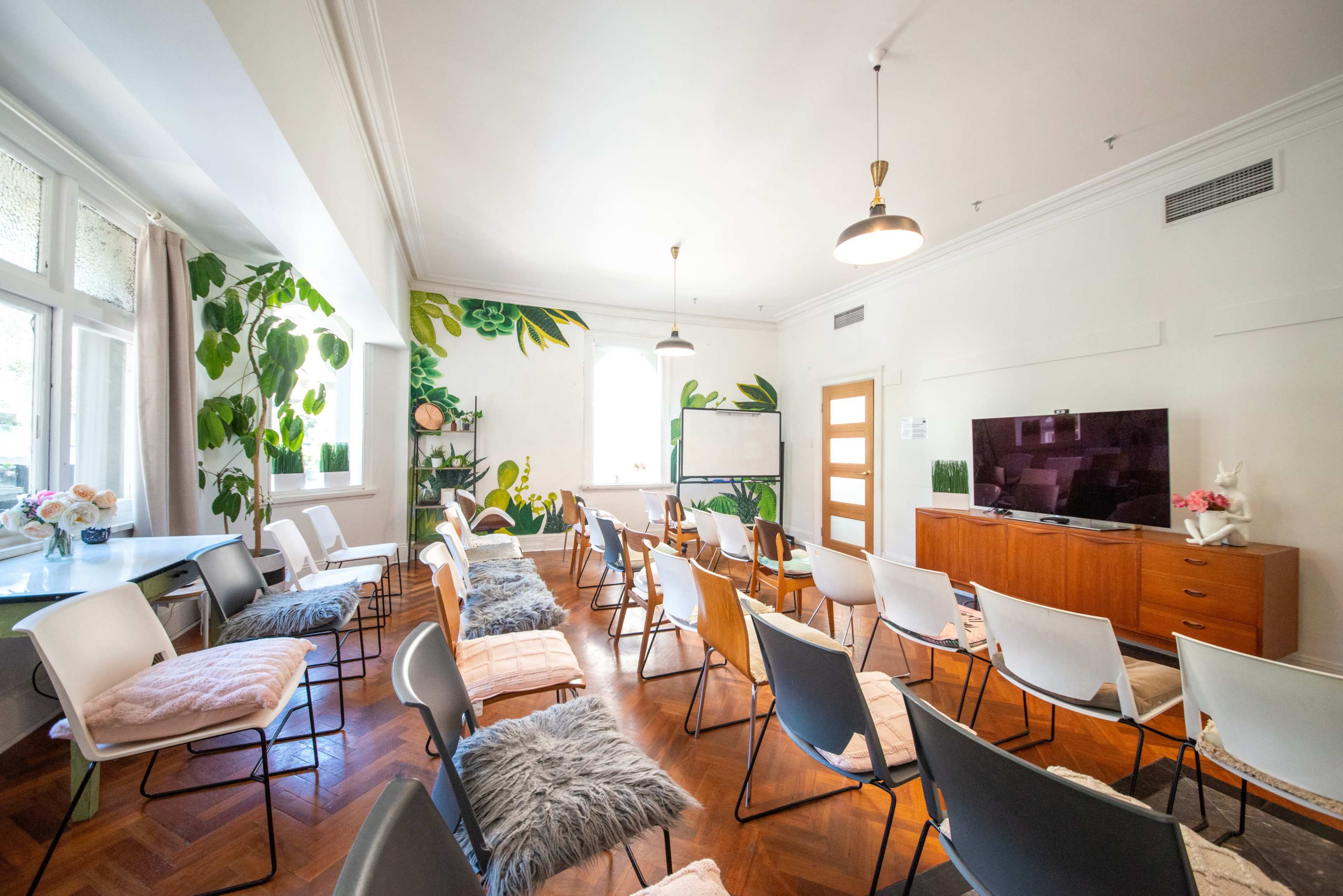 The room features arranged chairs and a large television, with plants painted on the walls and wooden accents throughout.