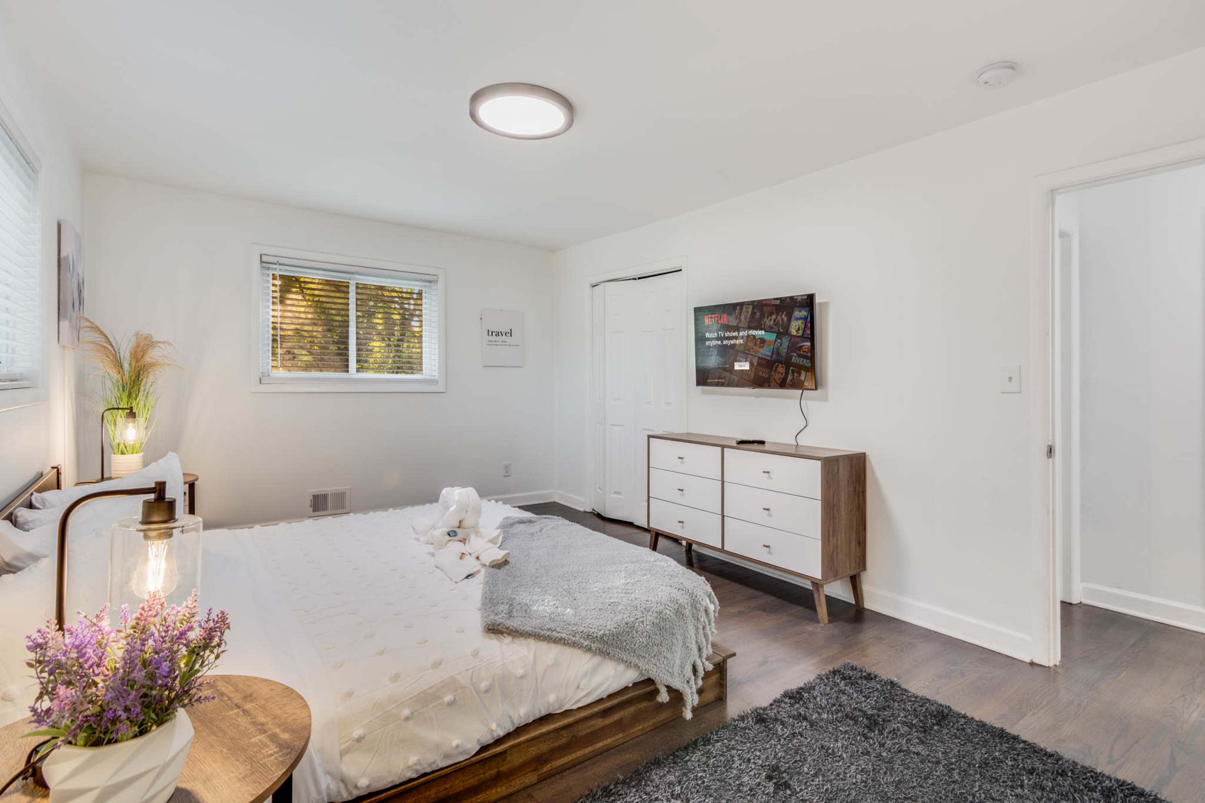 The image shows a well-lit bedroom featuring a bed with a light-colored blanket, a television mounted on the wall, and a dresser beside a window.