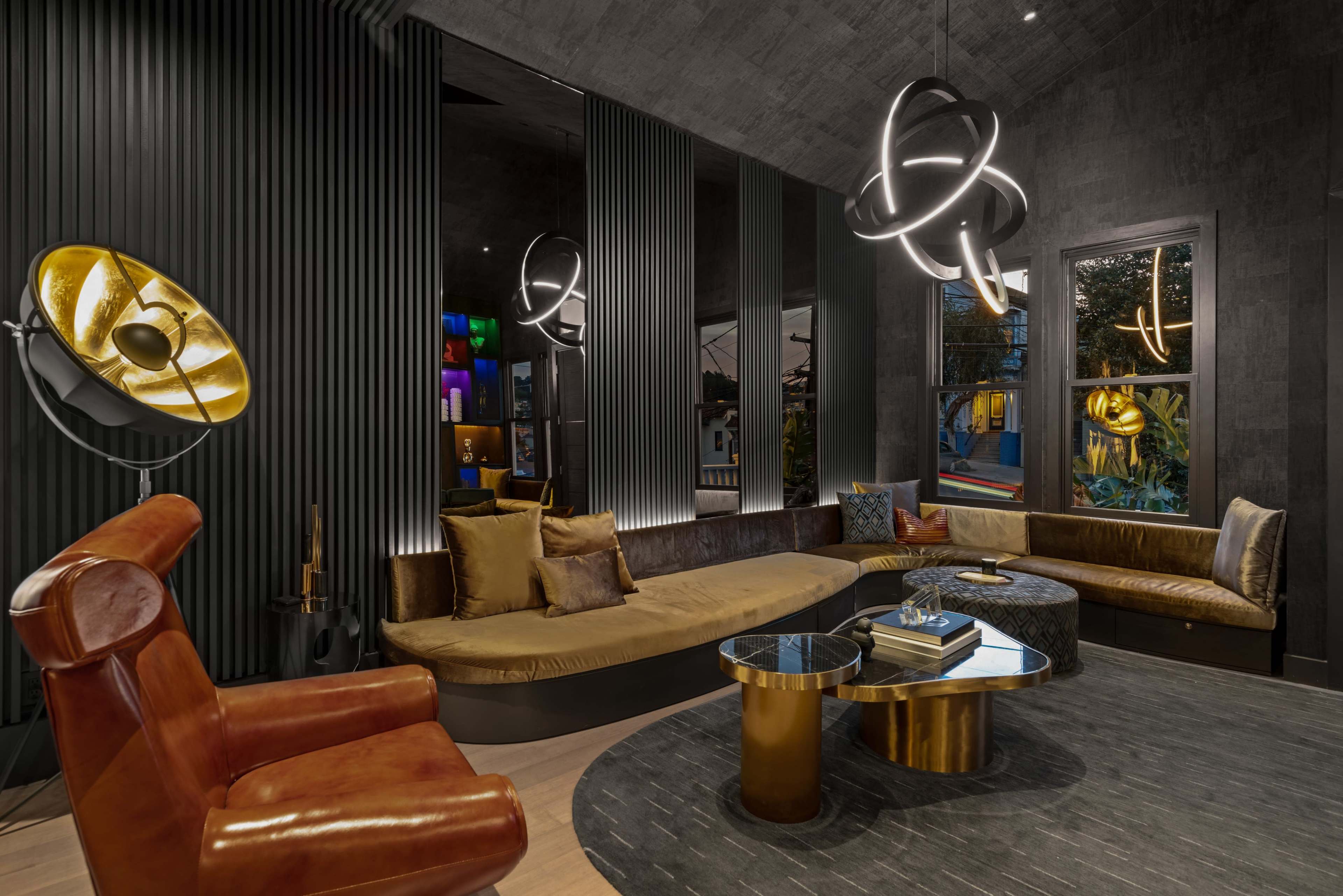 A modern lounge area featuring a circular sofa, a round table, a leather armchair, and stylish light fixtures against a backdrop of textured walls and large windows.
