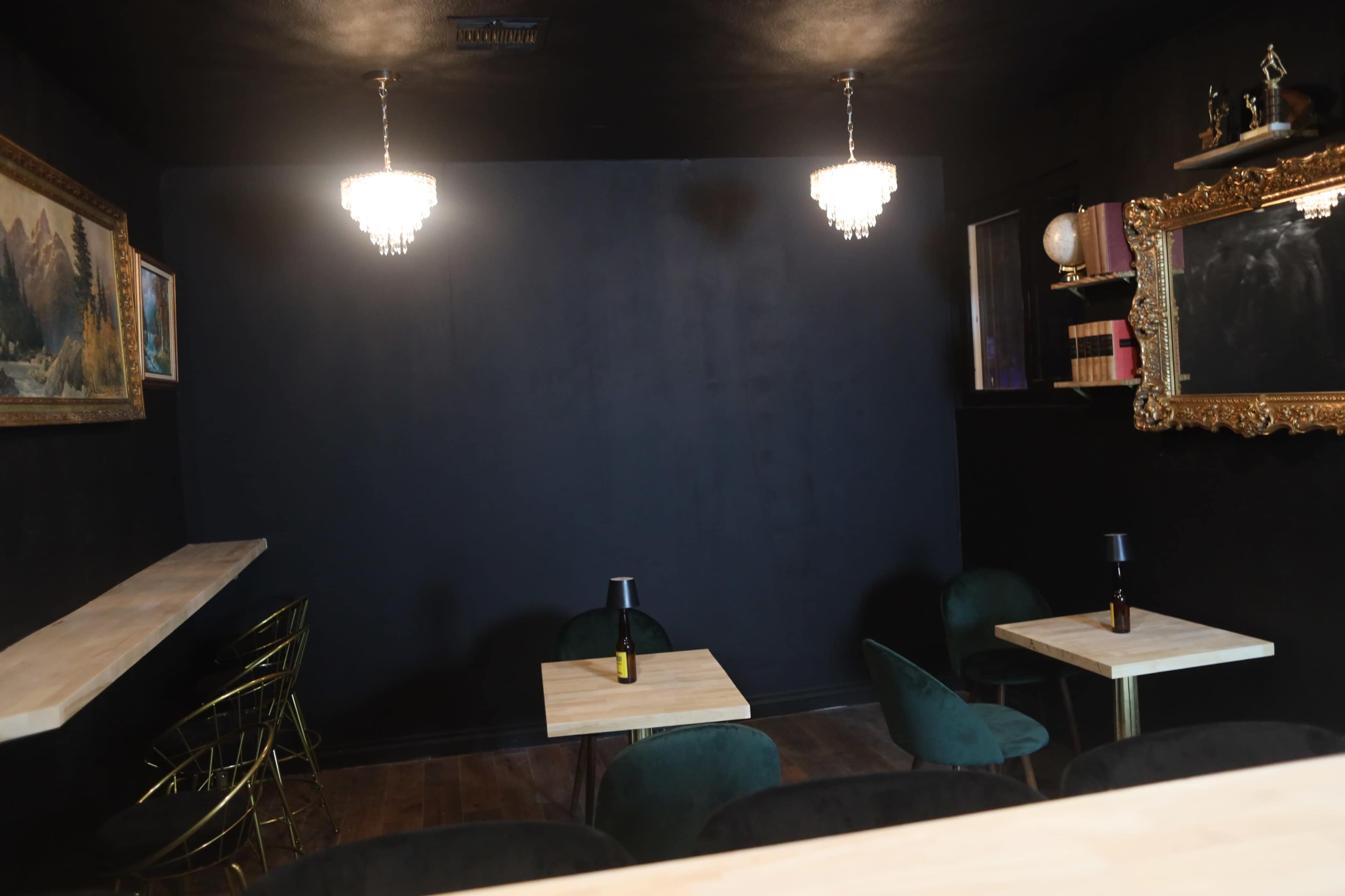 The image shows a dimly lit café with black walls, two decorative chandeliers, and tables with green chairs placed around a wooden counter.