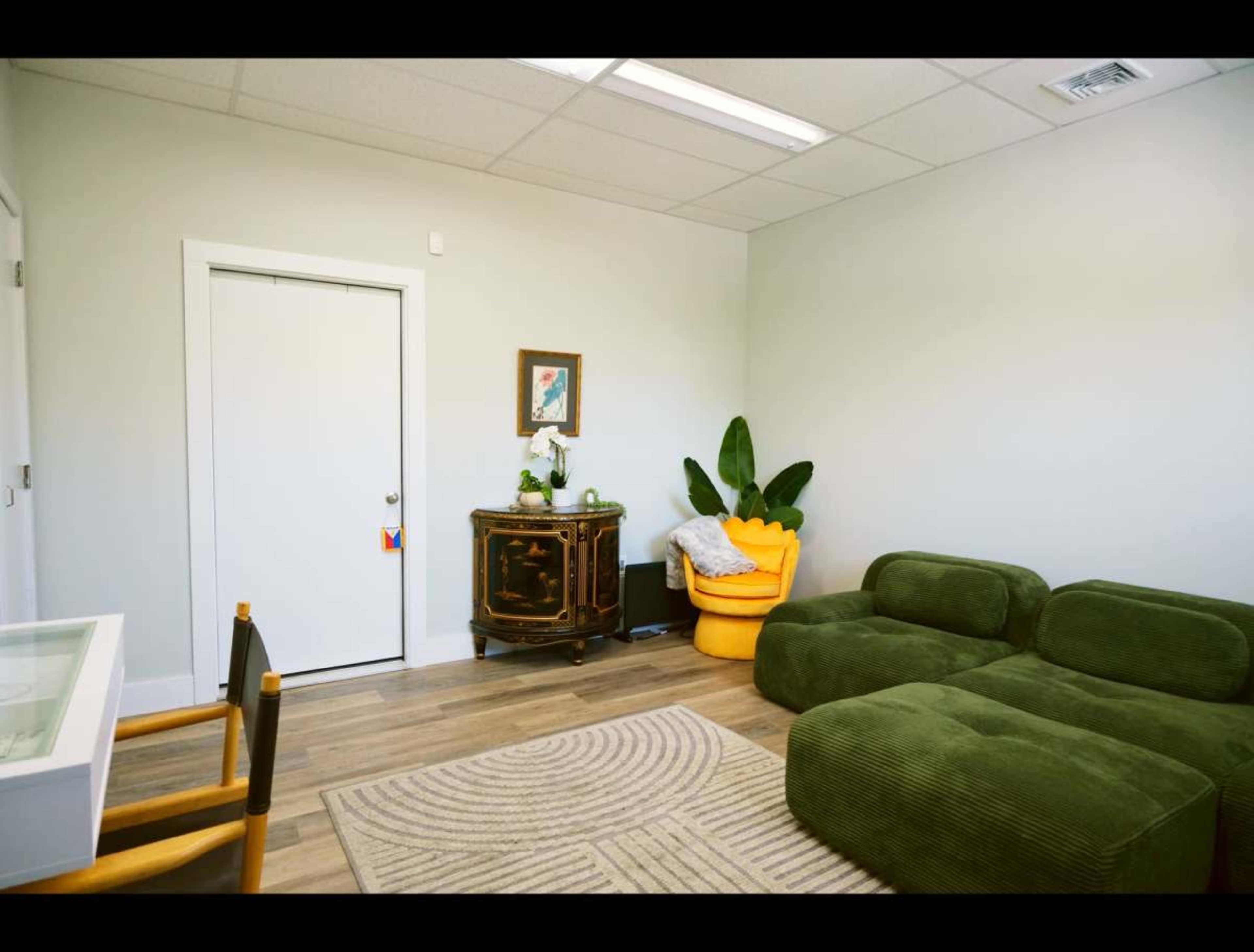 The image shows a modern, minimalist room featuring a green sofa, a yellow armchair, a decorative cabinet, and a door leading to another space.