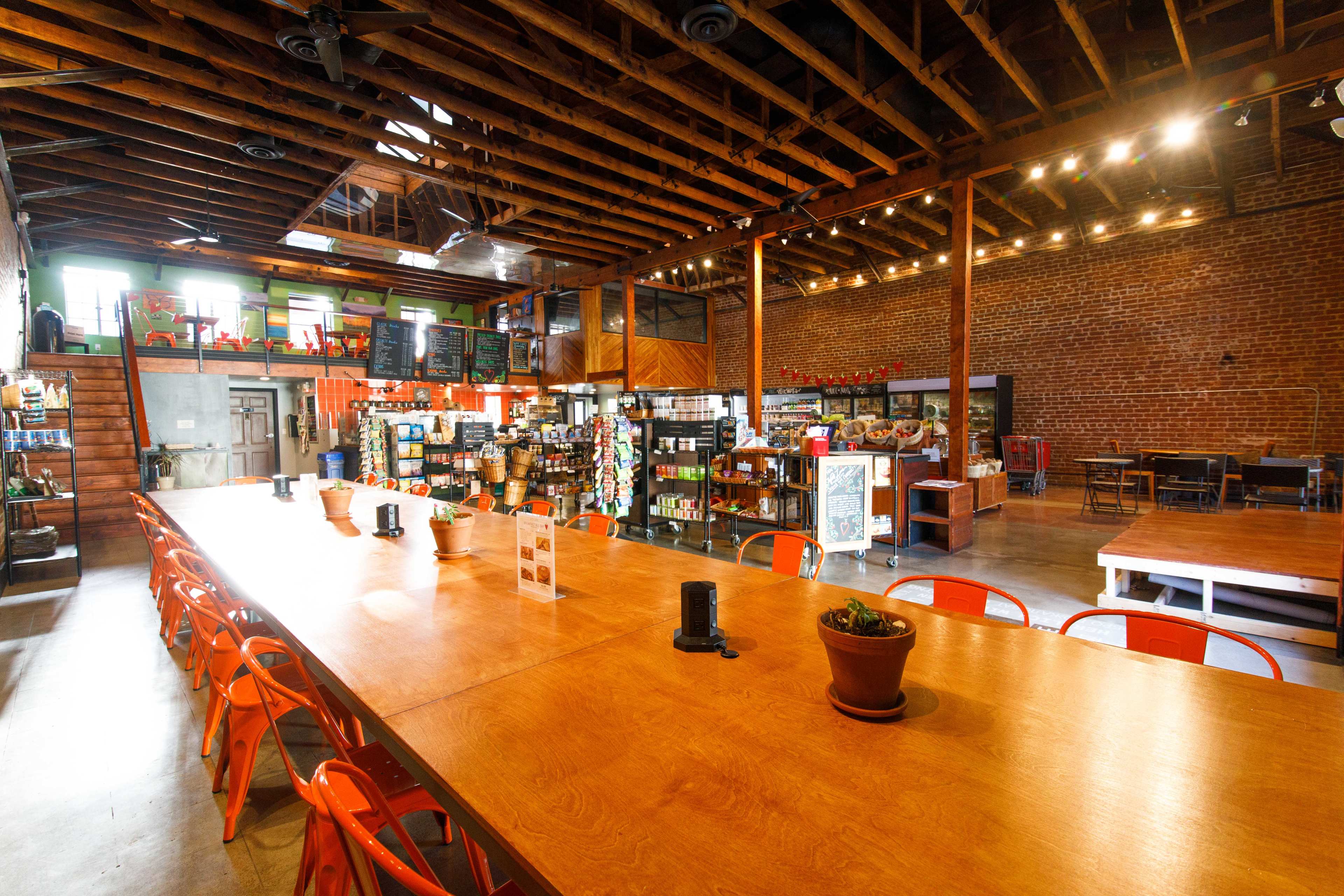 The image shows a spacious café with a long wooden table, bright orange chairs, exposed brick walls, and hanging lights.
