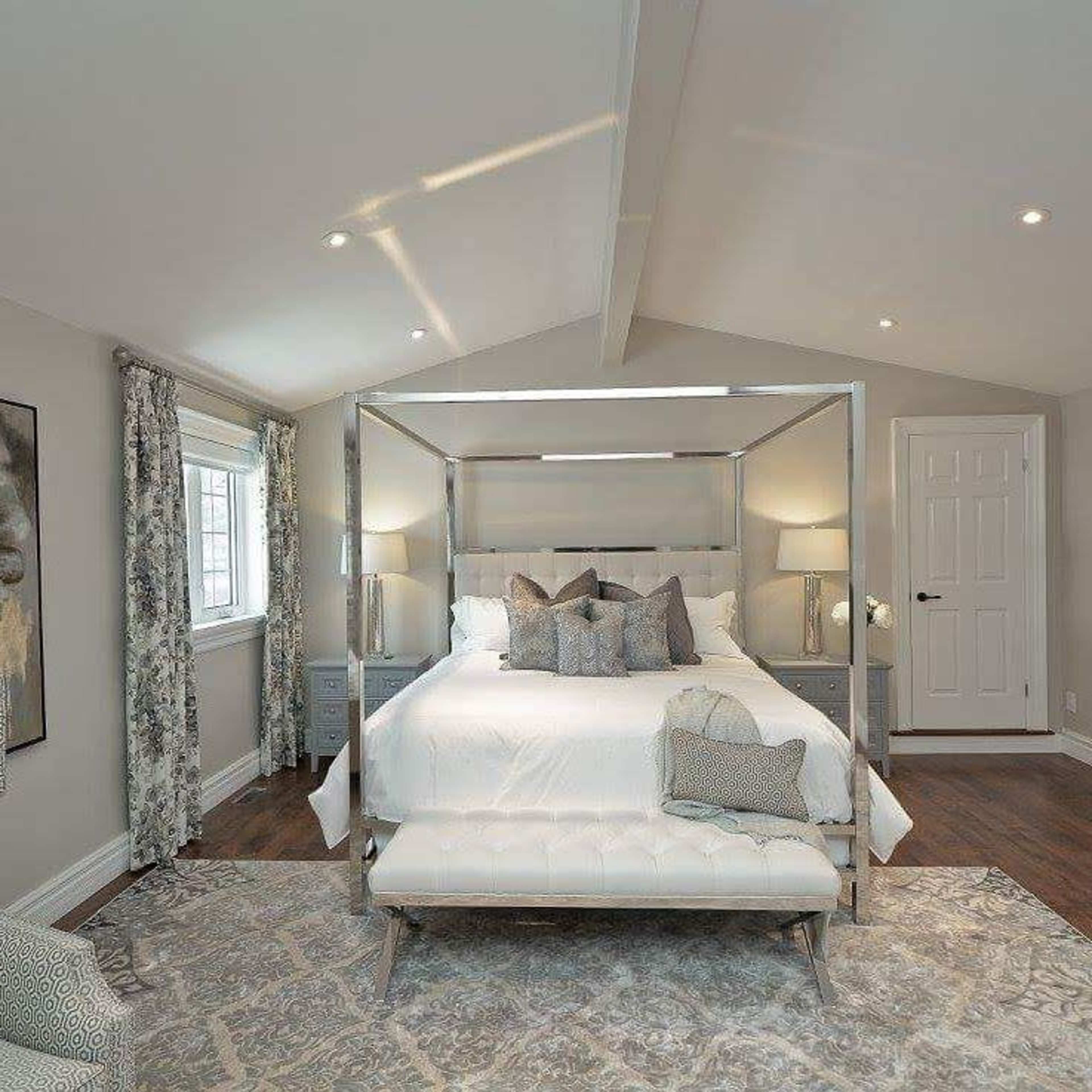 A modern bedroom featuring a four-poster bed, a cozy bench at the foot of the bed, and large windows with patterned curtains.
