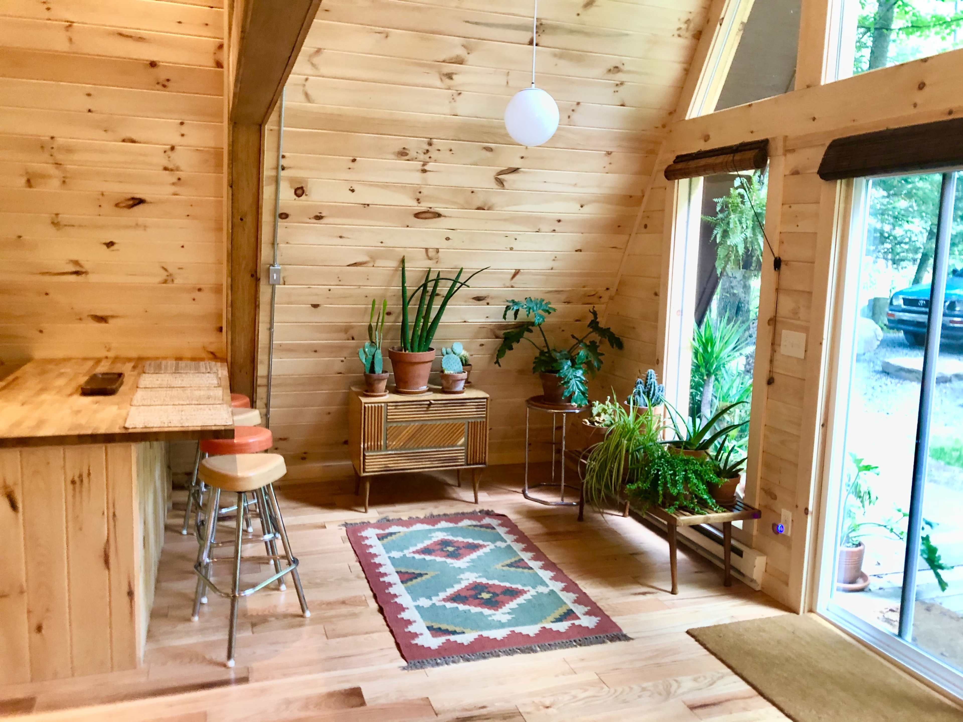 The image shows a wooden interior featuring an A-frame design, with a small dining area, a rug with geometric patterns, potted plants, and large windows providing natural light.