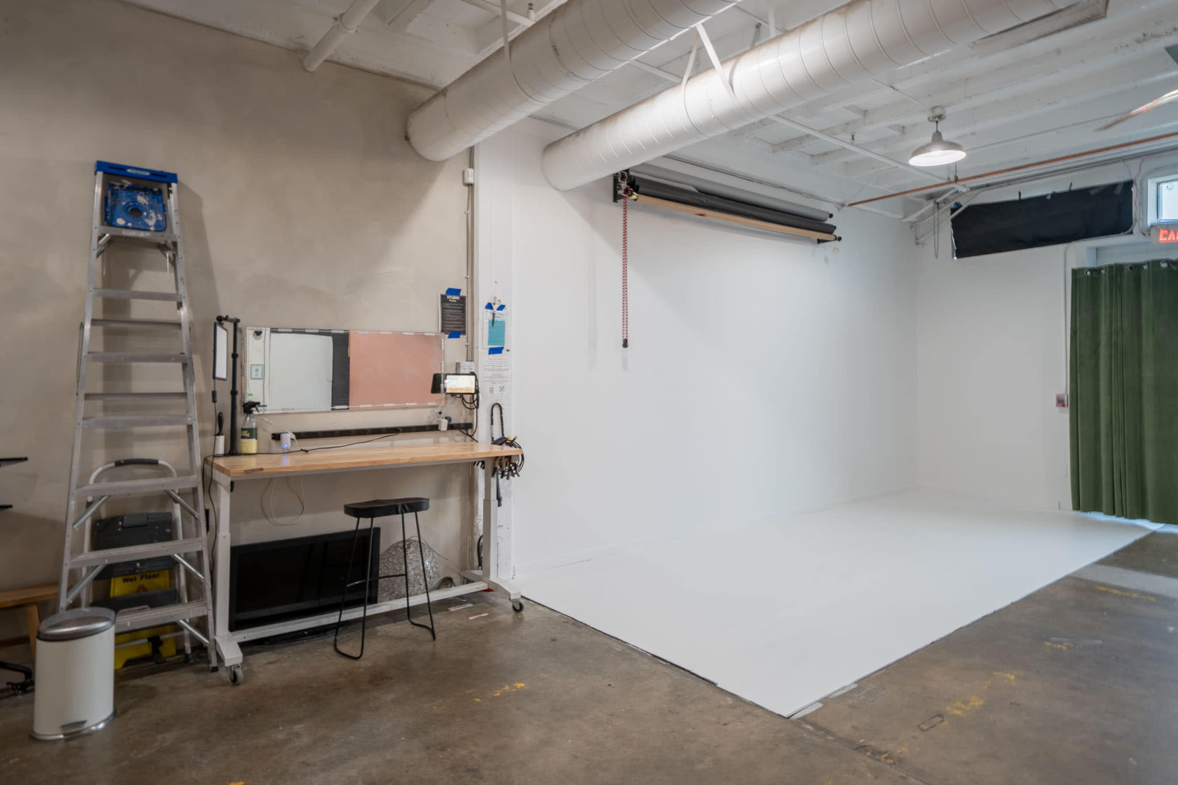 The image shows a spacious, well-lit studio with a white wall and a roll of backdrop material, accompanied by a ladder, workbench, and black stool.