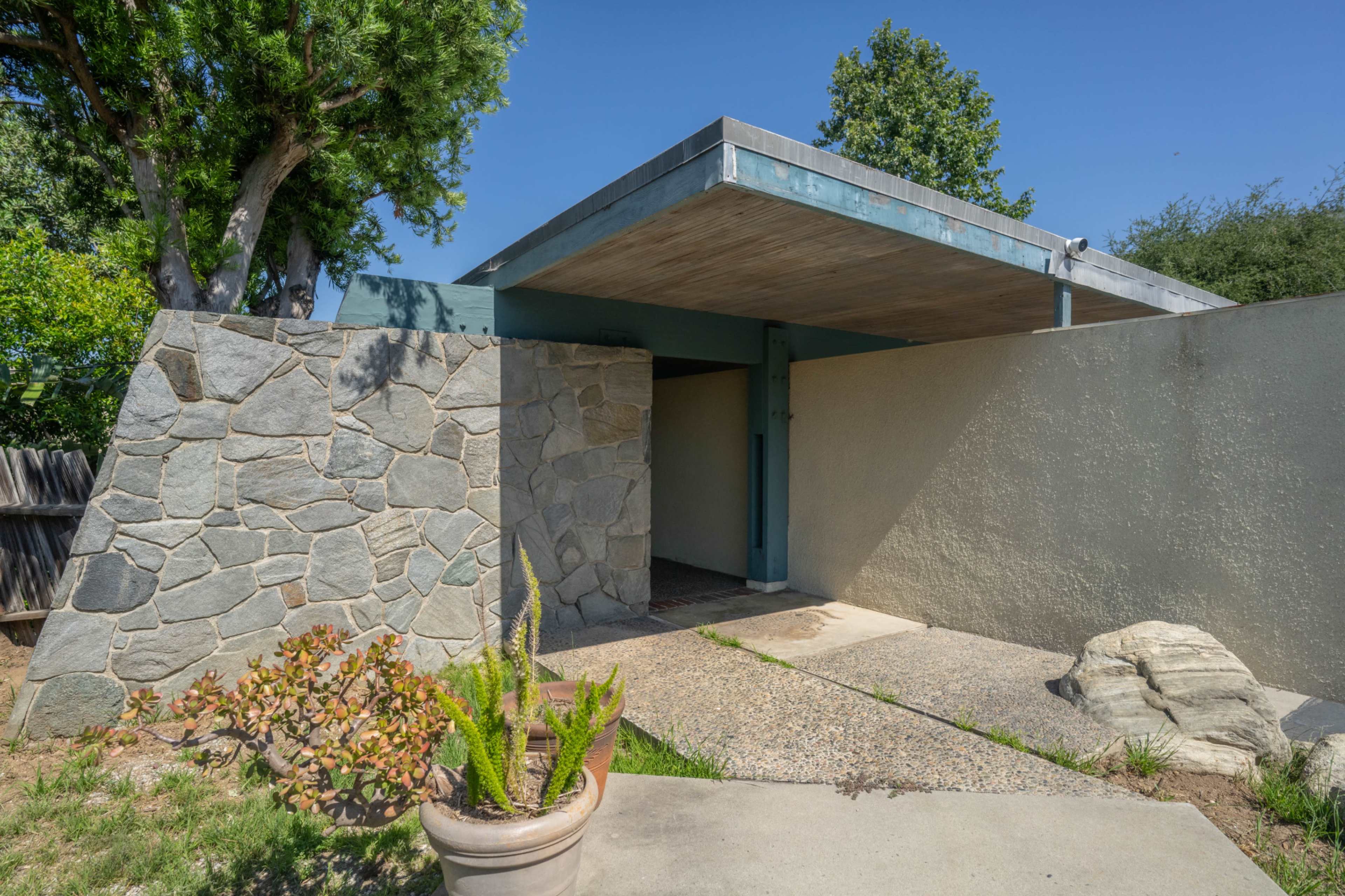 The image shows a modern architectural entrance with a flat roof, stone walls, and a pathway leading into the building.