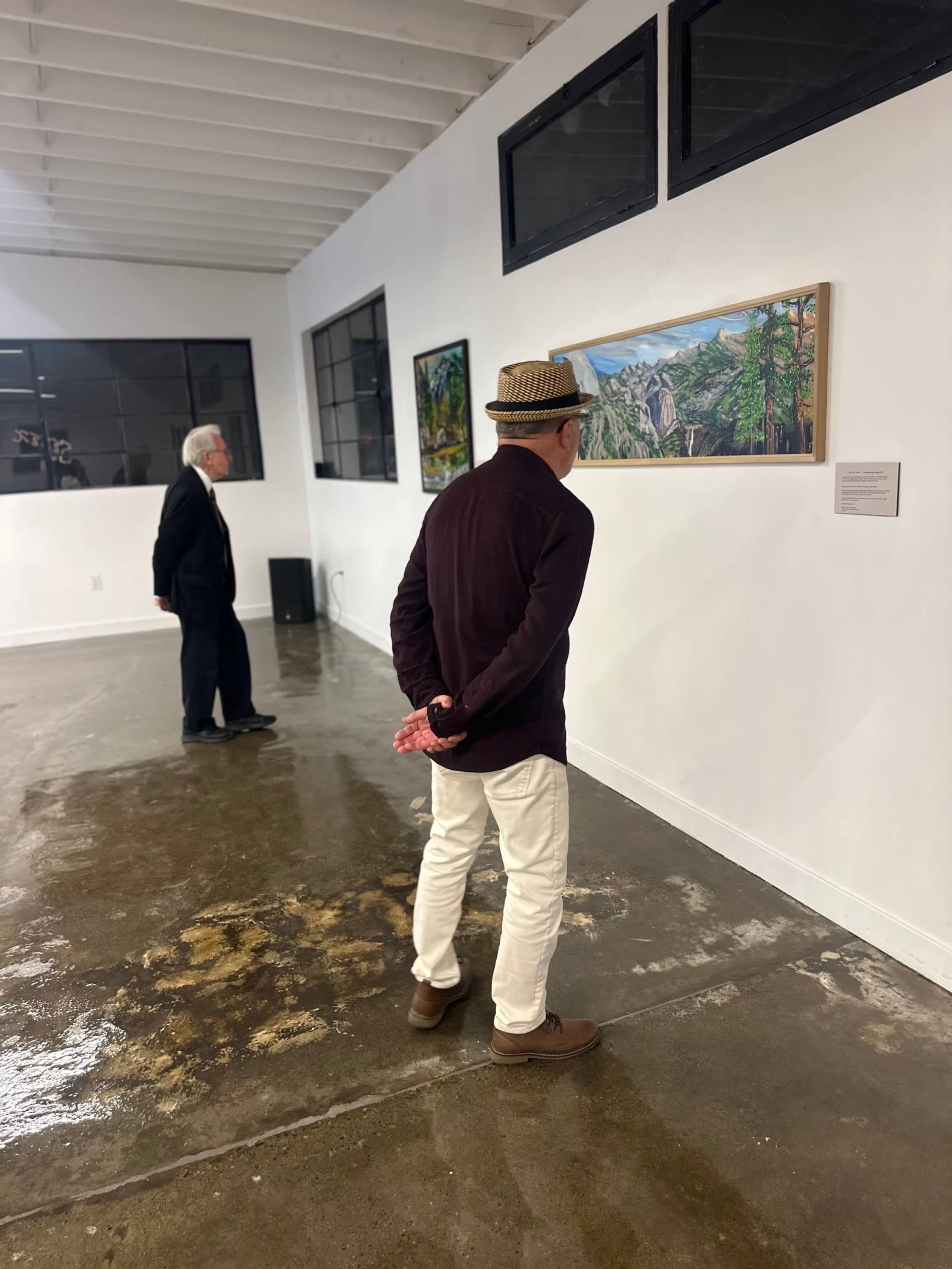 Two men are standing in an art gallery, with one looking at a painting while the other stands nearby.