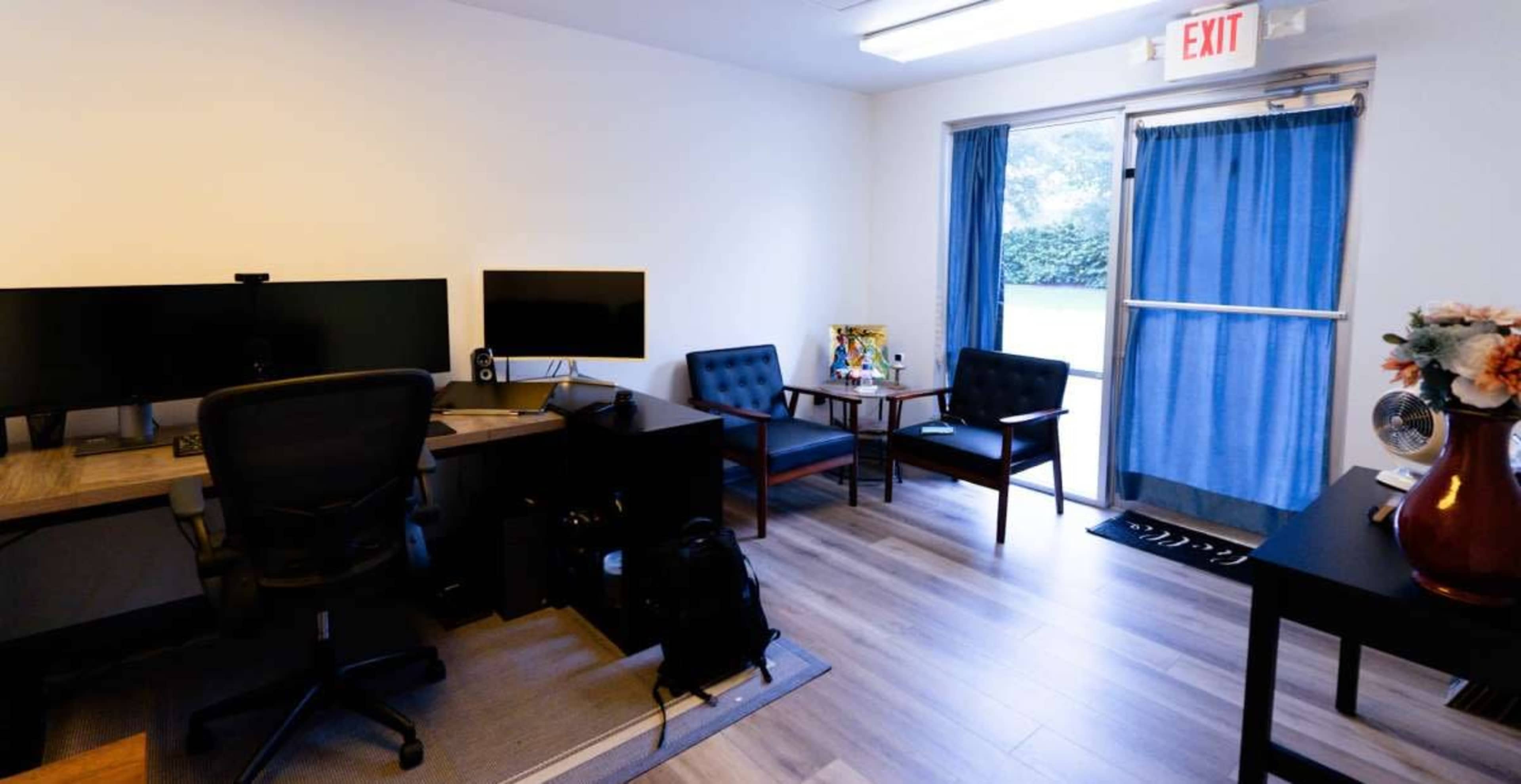 The image shows a modern office space featuring two computer monitors on a desk, a black swivel chair, two armchairs with a small table between them, and an exit door with blue curtains.