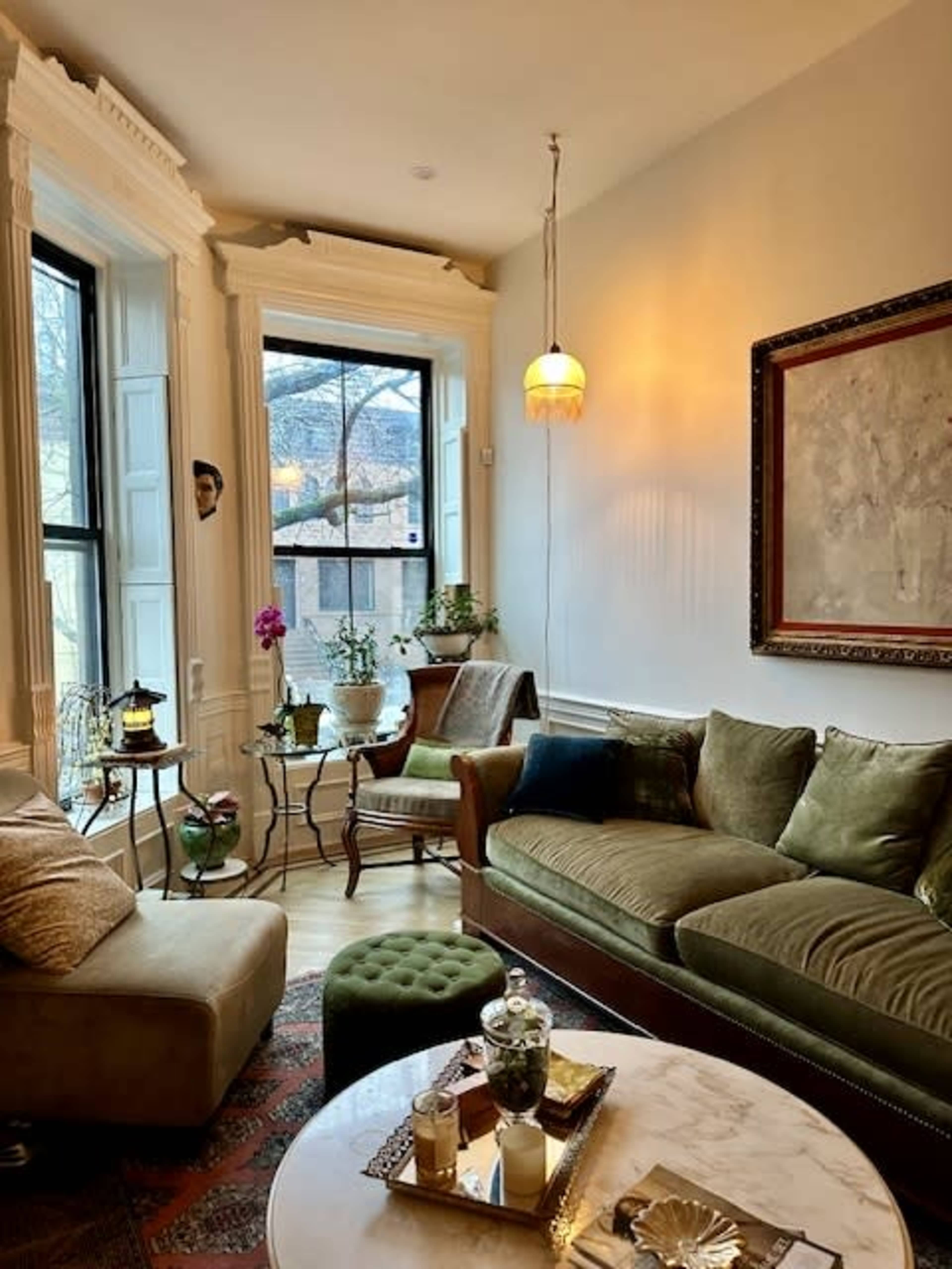 The image shows a cozy living room with a green velvet sofa, a small table with decorative items, and a window overlooking a street.