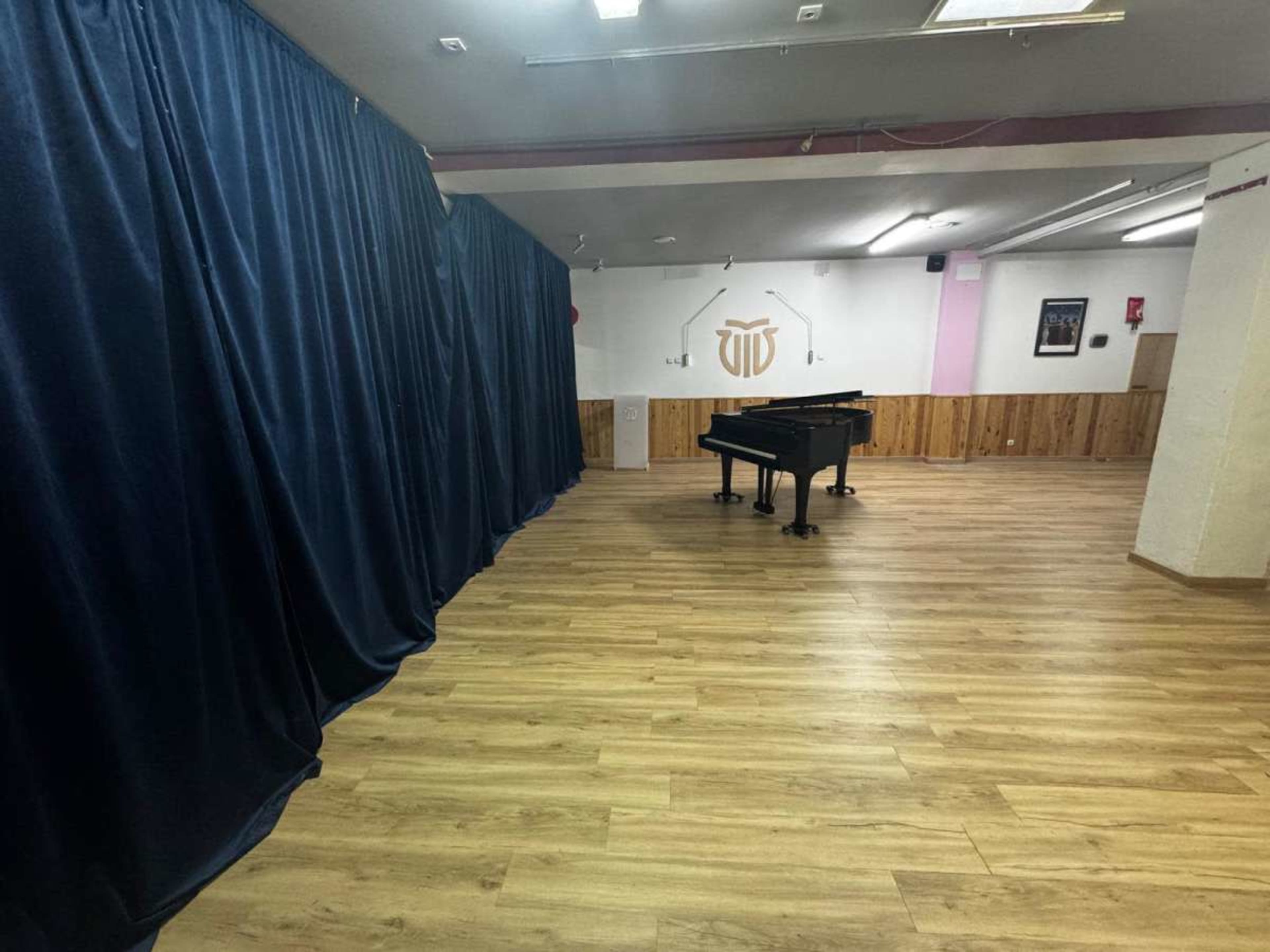 The image shows a spacious room with wooden floors, featuring a grand piano in the center and dark blue curtains along one wall.