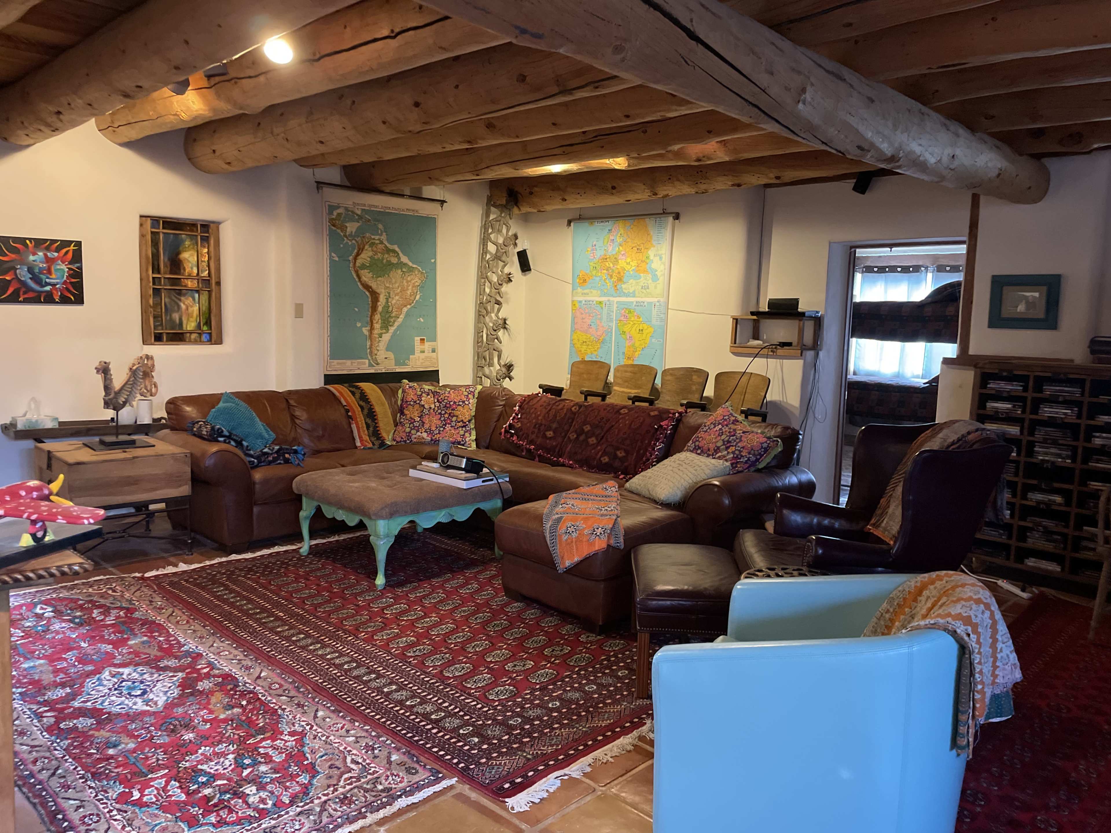 The living room features a large brown sectional sofa with various patterned throws, surrounded by vintage maps, rugs, and rustic wooden beams.