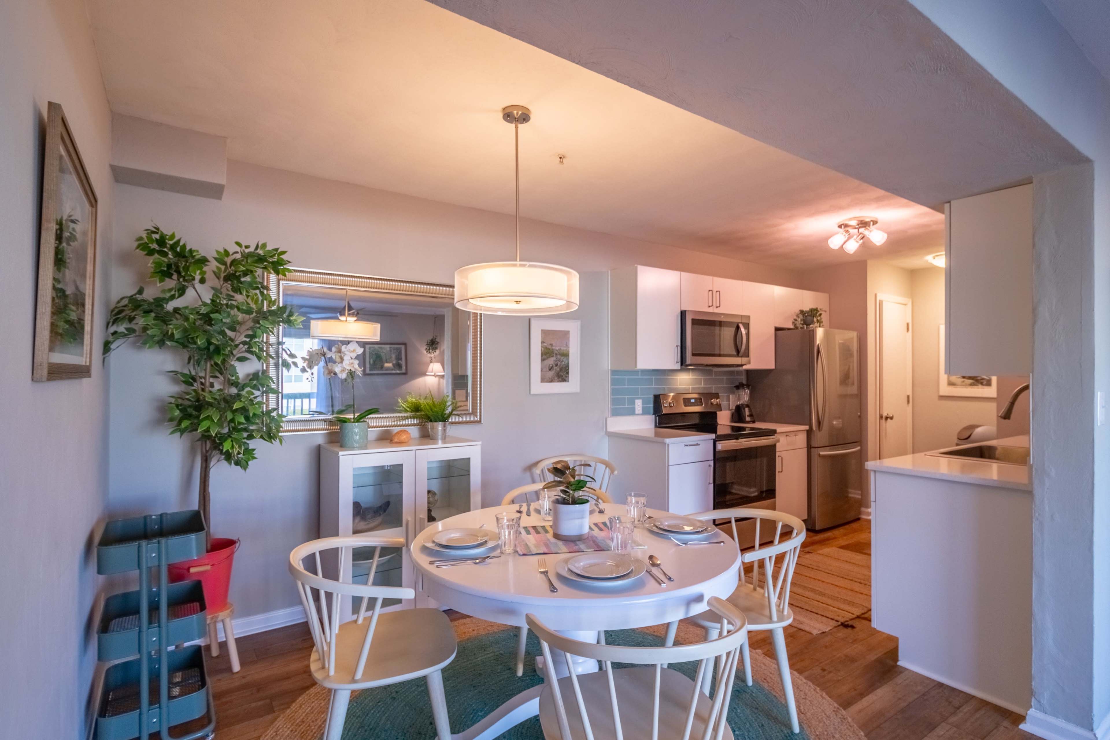 A modern kitchen-dining area features a round white table set for four, surrounded by chairs, with a potted plant and a mirror on the wall.