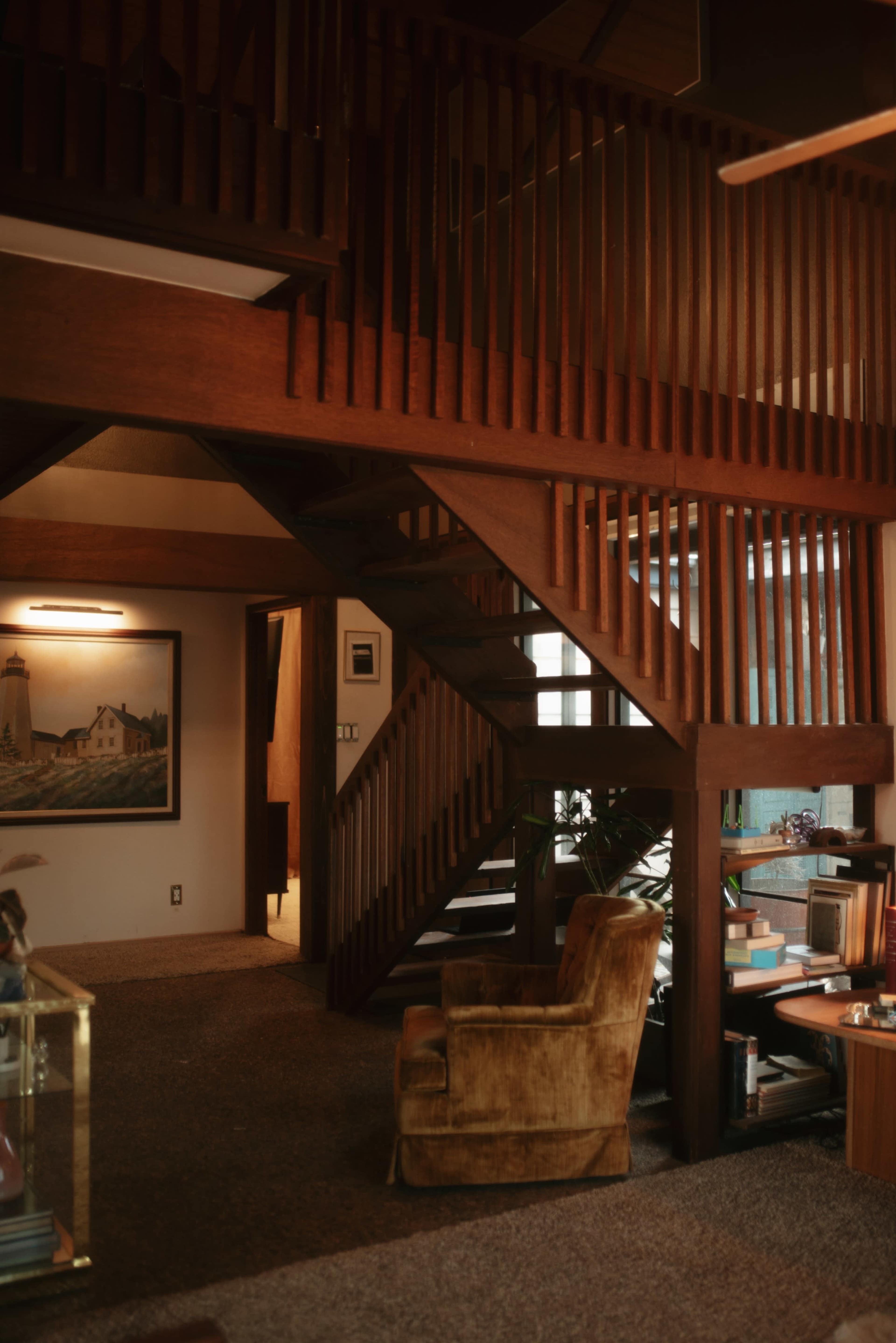 The image shows a wooden staircase leading to an upper level in a cozy interior space, accompanied by a chair and framed artwork on the walls.