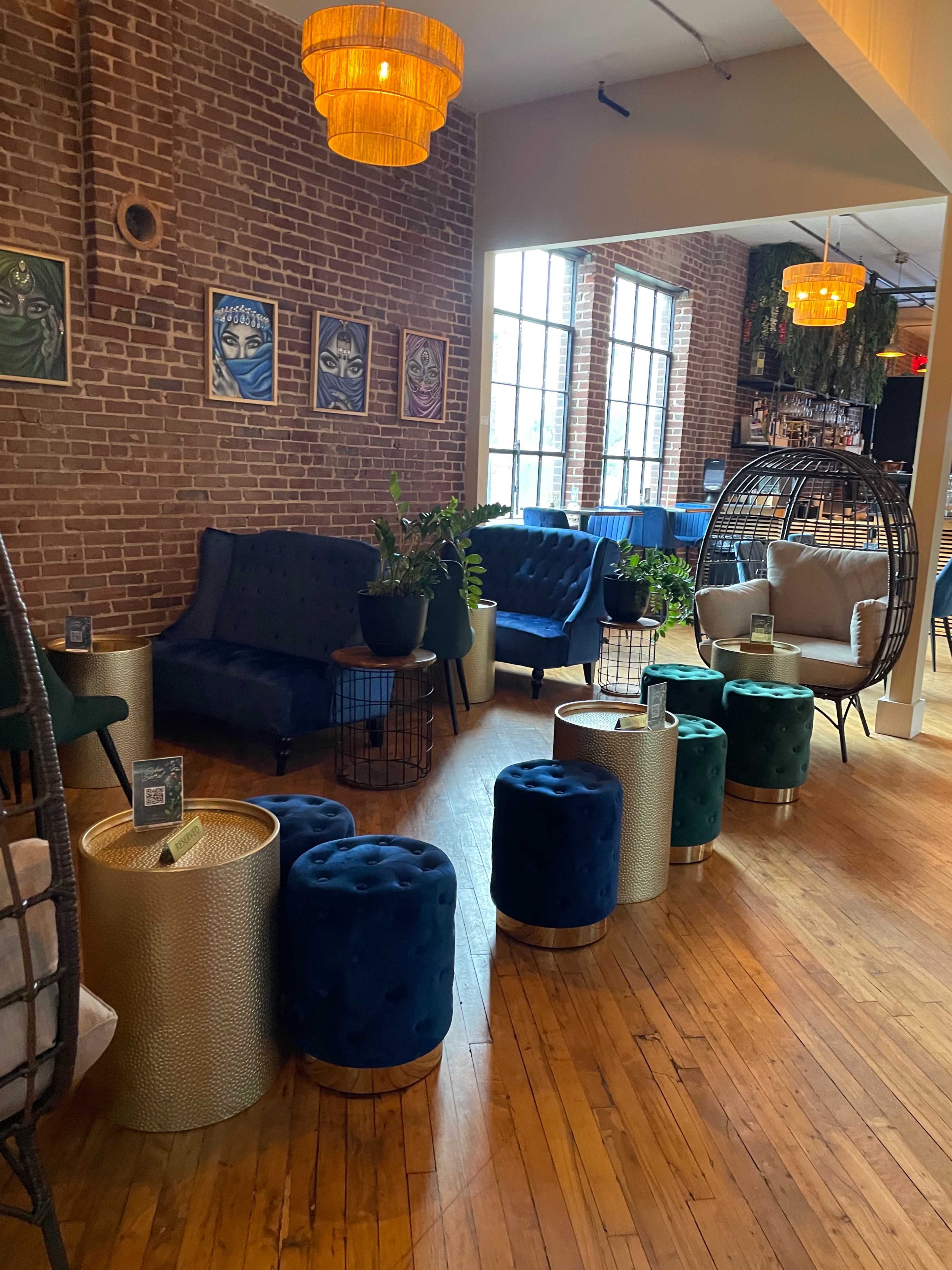 A cozy lounge area with brick walls, various seating options including plush sofas and woven hanging chairs, and decorative plants arranged around small side tables.