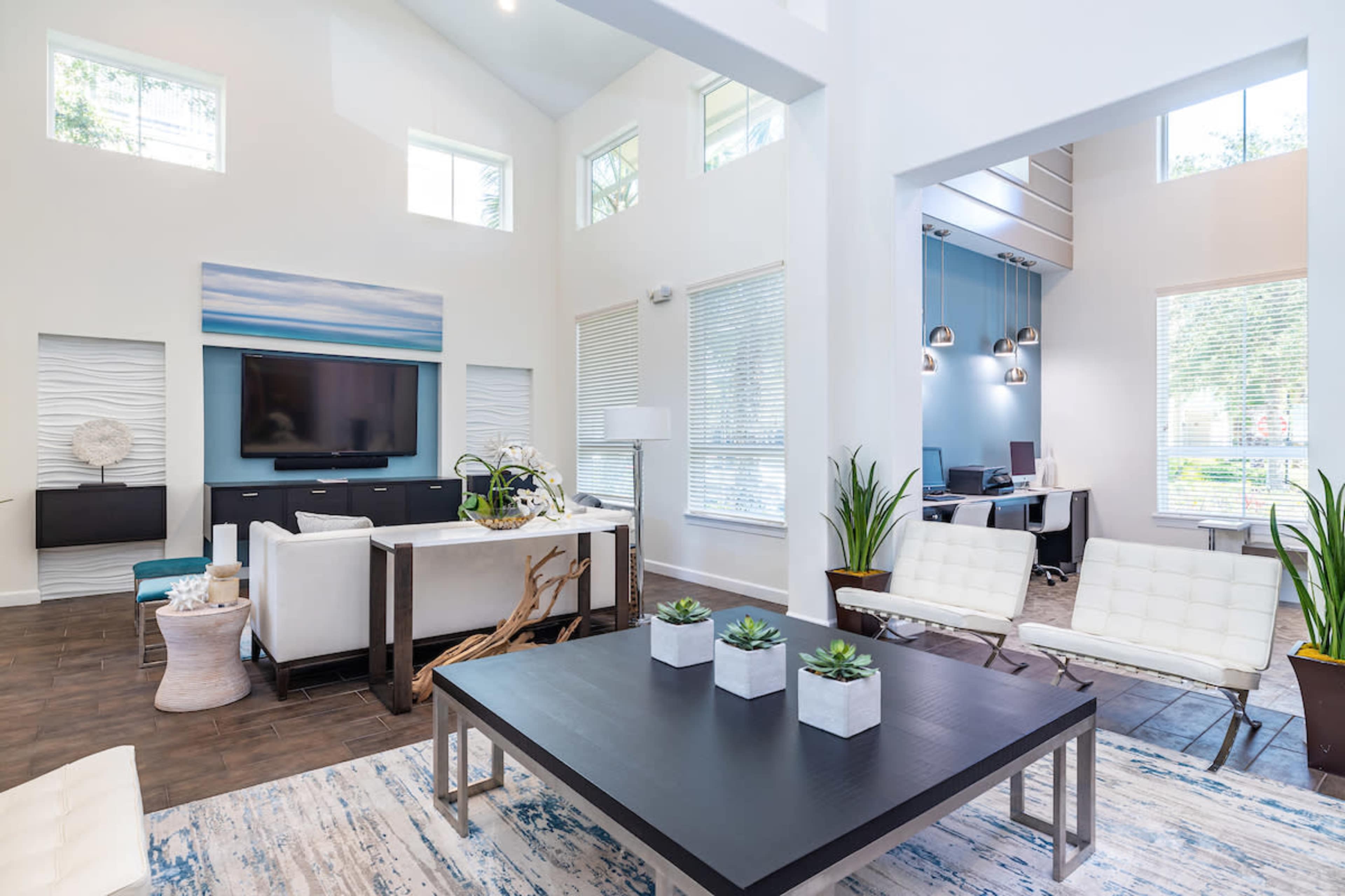 A modern living room with a large flat-screen TV, a coffee table, white furniture, and an adjacent workspace with desks.