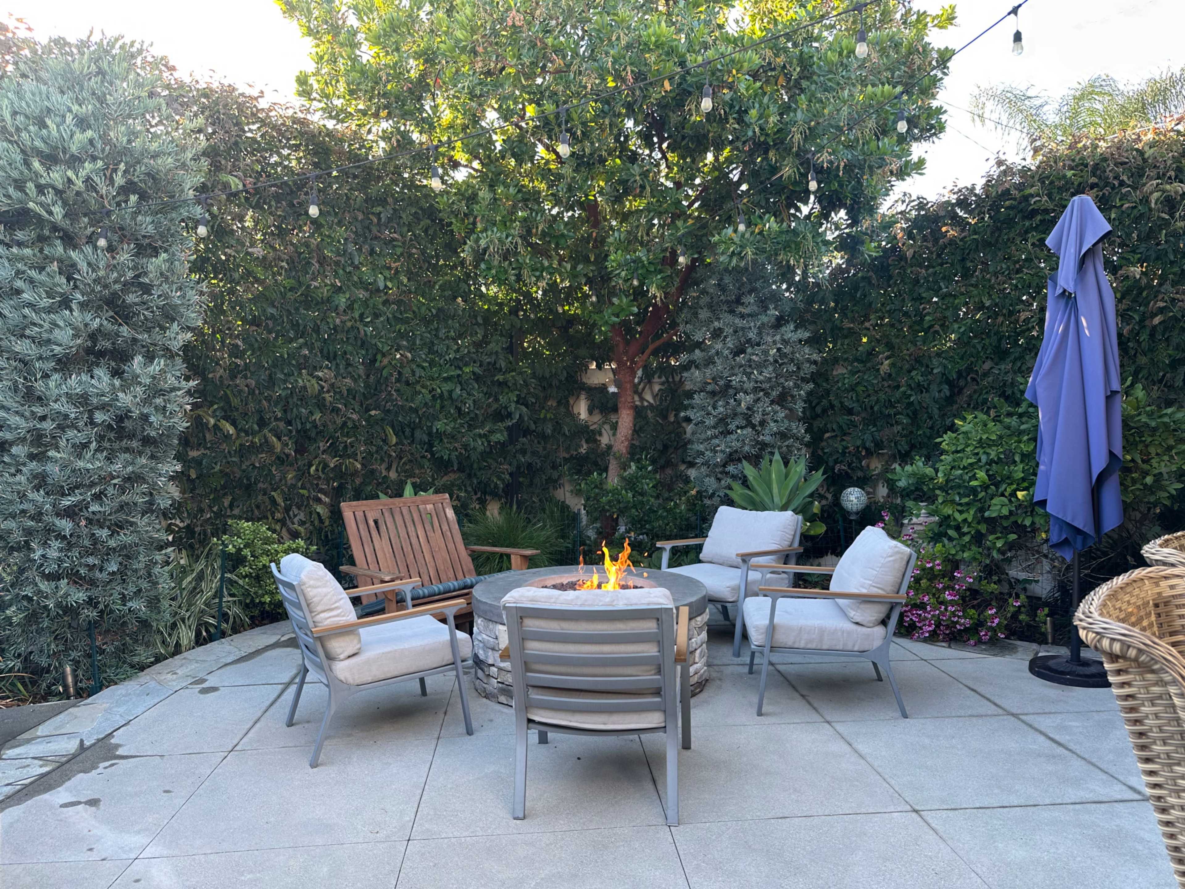 A patio features a circular fire pit surrounded by four chairs, with lush greenery and a large umbrella in the background.
