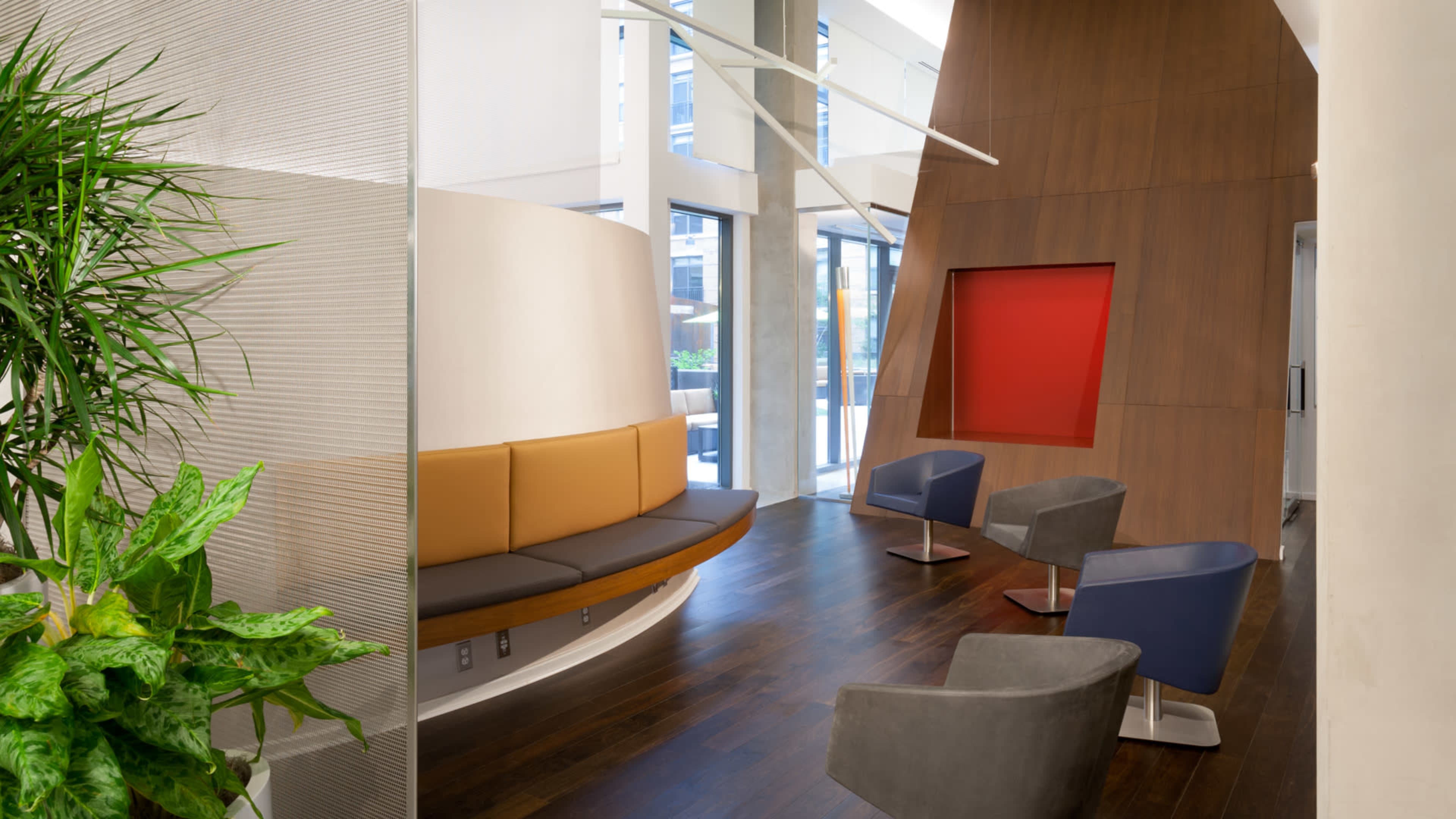 A modern interior waiting area with a curved bench, several chairs, and a large window allowing natural light to enter.