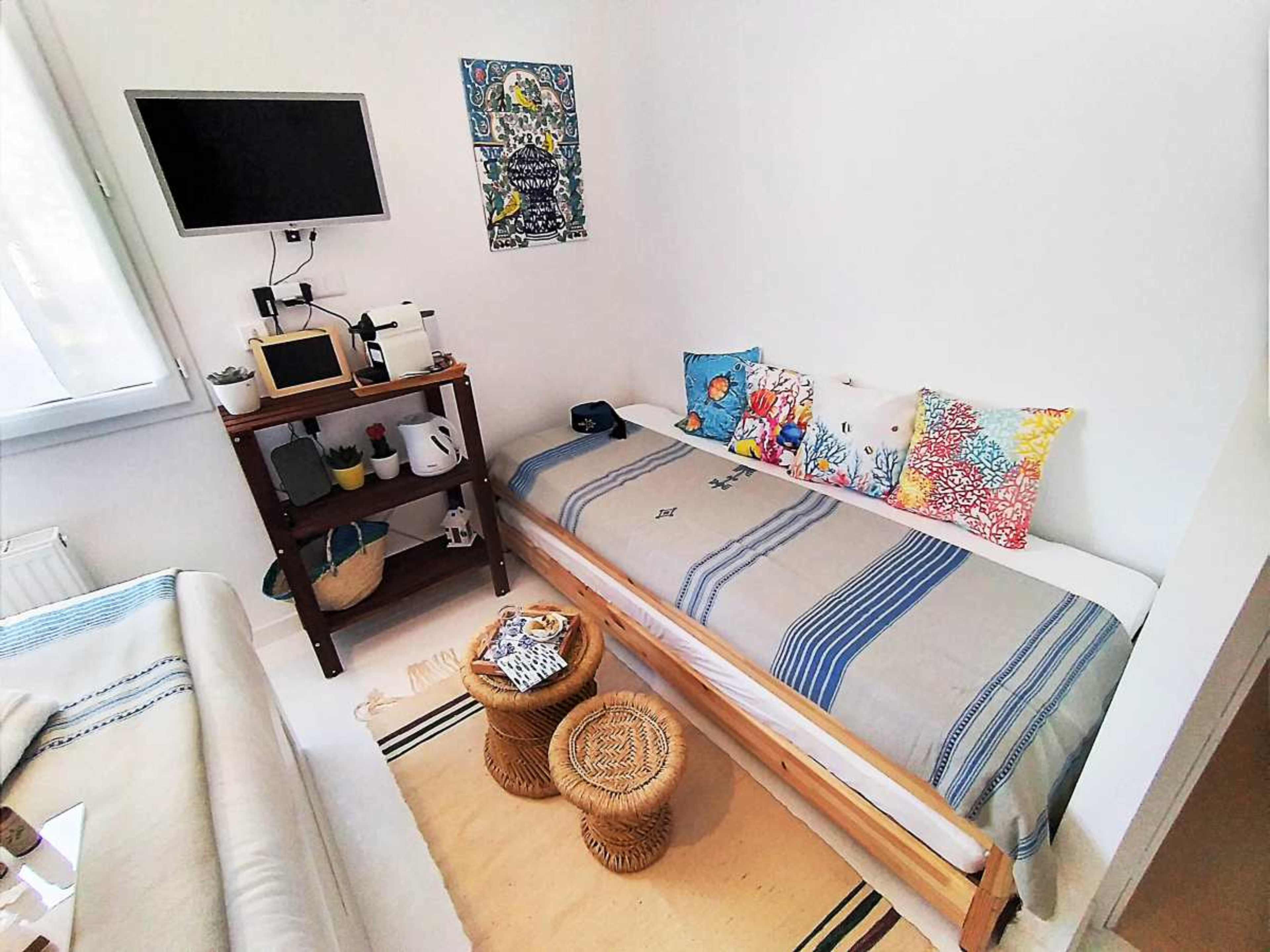 A small, minimalist bedroom featuring a single bed with decorative pillows, a wall-mounted TV, a wooden shelf with various items, and a light-colored rug on the floor.