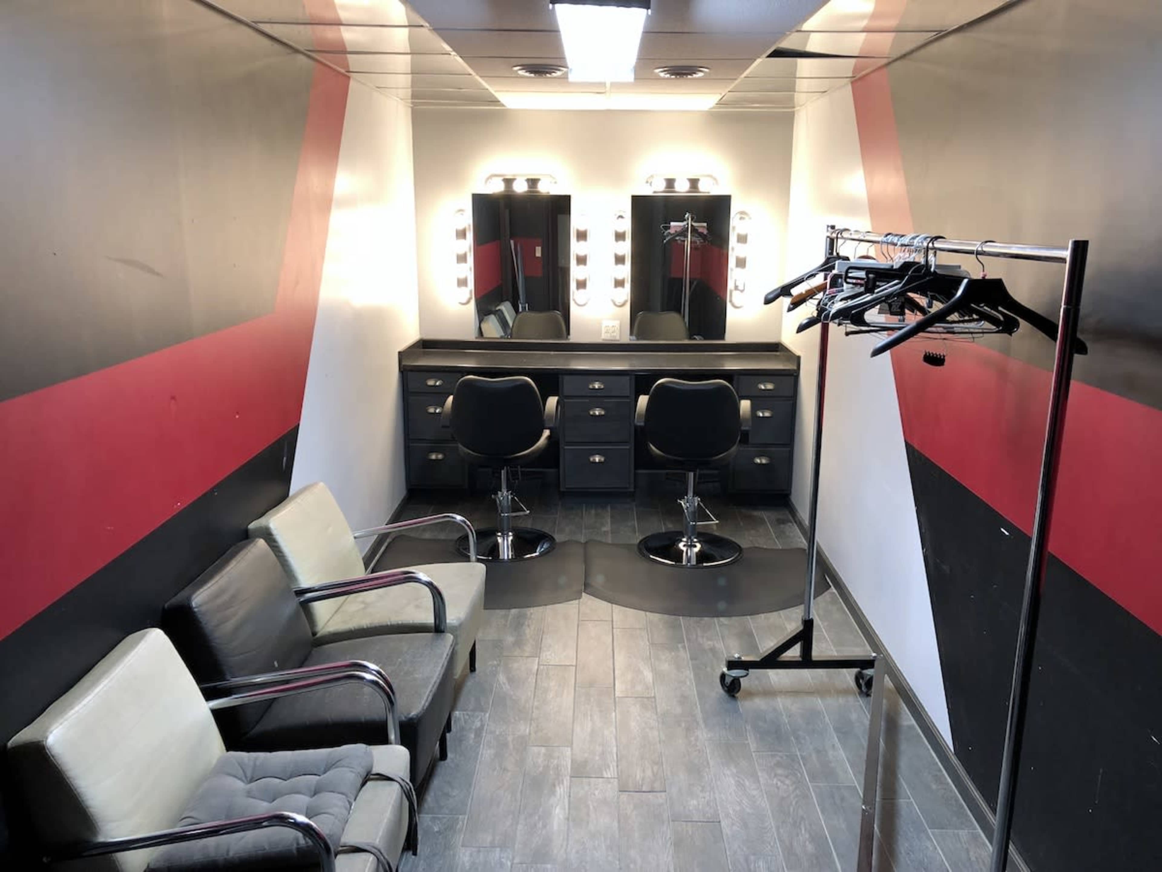 The image depicts a moderately lit dressing room with two black chairs and a rack of clothes against a wall featuring red and black designs.
