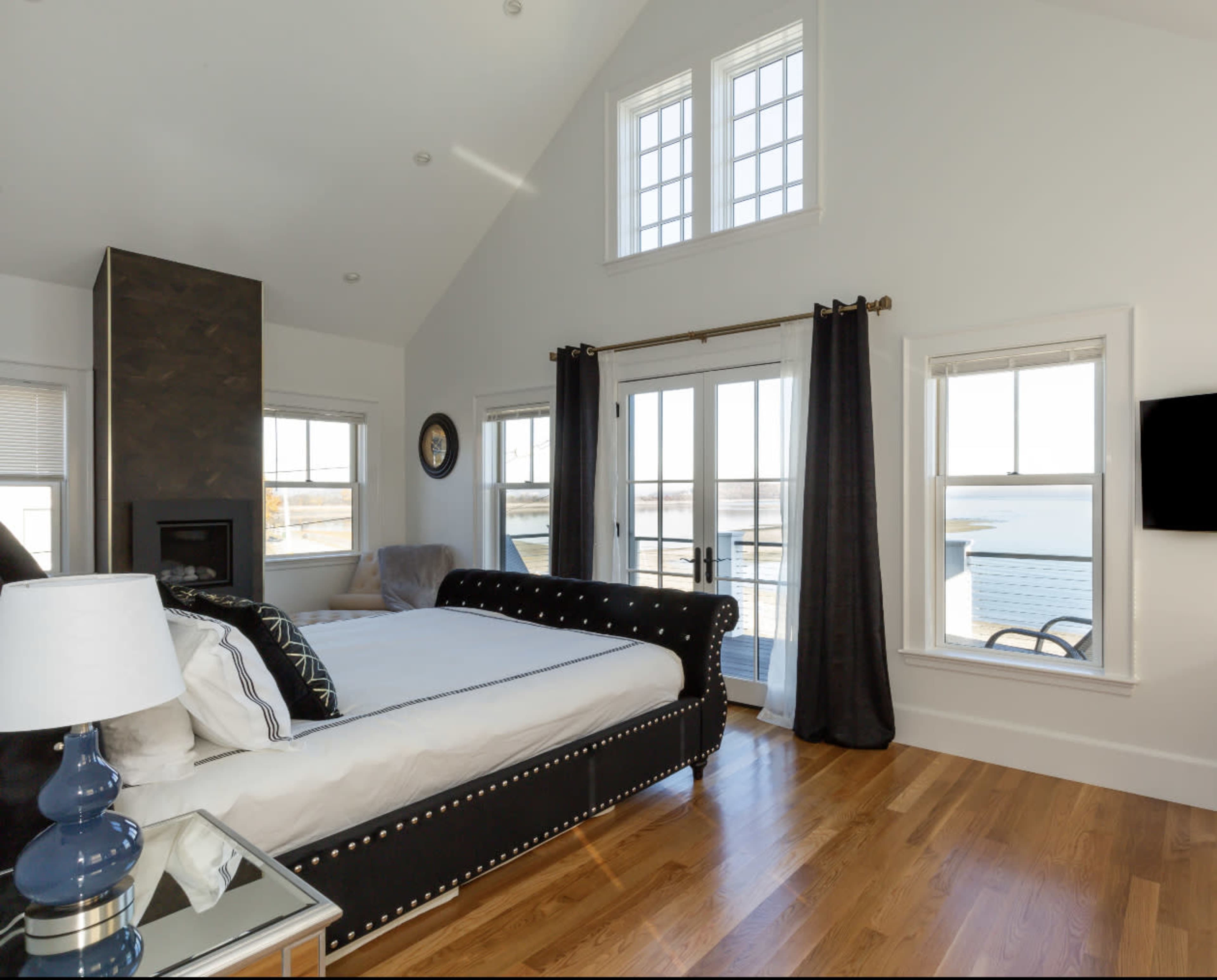 The room features a large bed with a black upholstered headboard, multiple windows allowing natural light, and a pair of French doors leading to a view of the water.