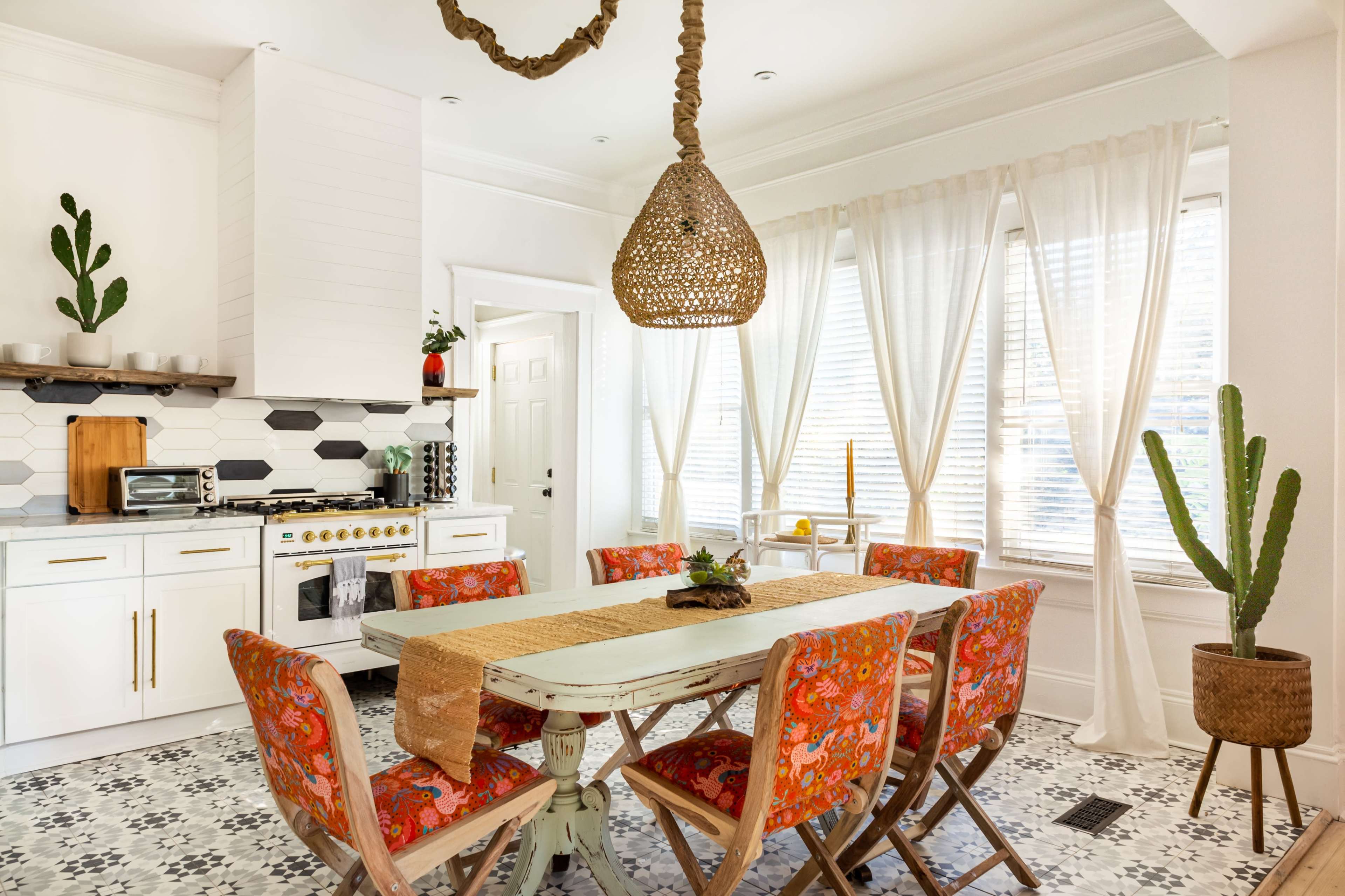A bright kitchen features a wooden dining table surrounded by colorful chairs, with a large pendant light hanging above and plants in the corners.