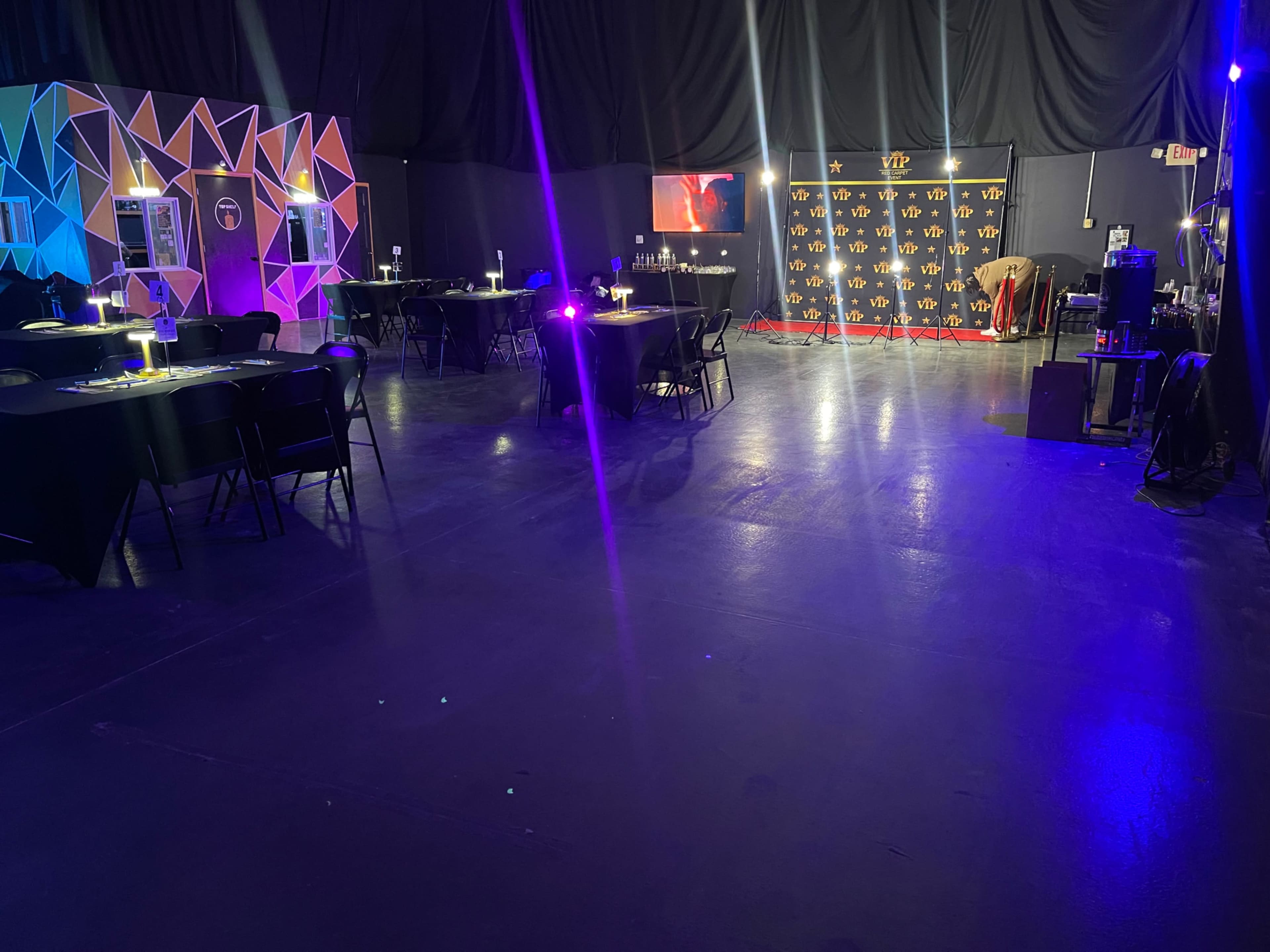 An event space with black tables and chairs, colorful geometric decorations on one wall, and a lit backdrop with a logo in the distance.