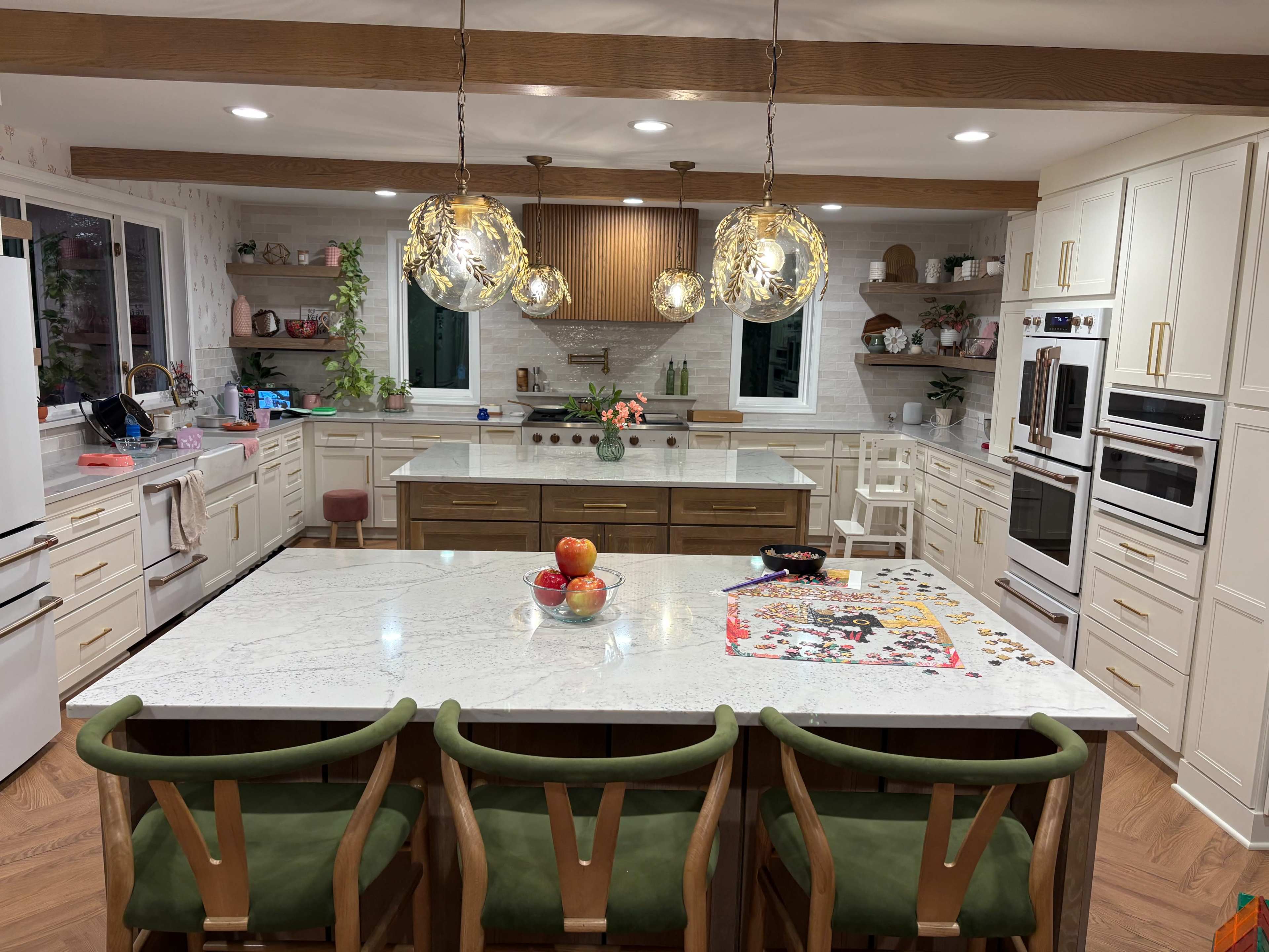 A modern kitchen features a large marble island with bar stools, pendant lighting, and open shelving displaying various kitchen items.
