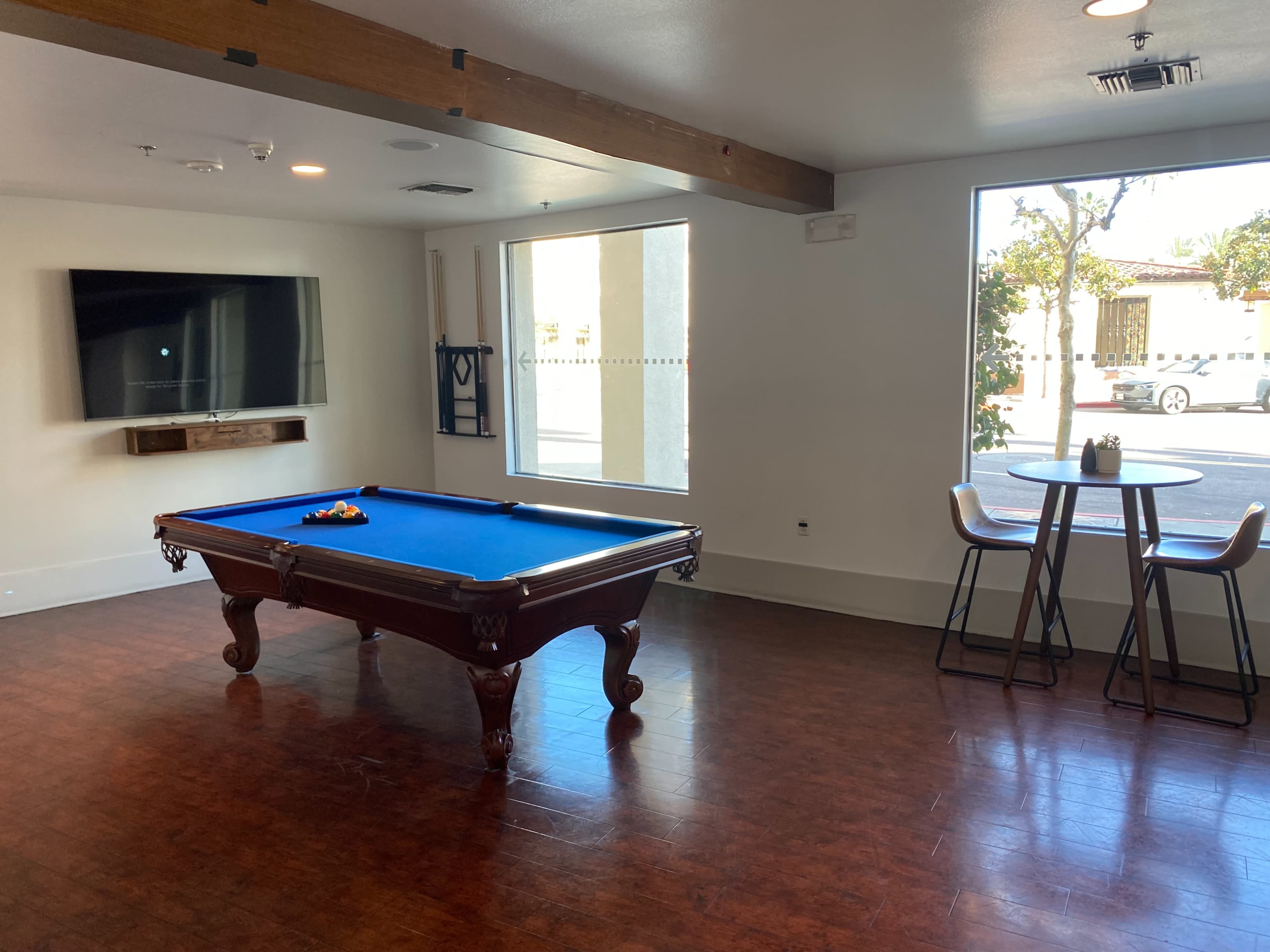 The image shows a room with a blue felt pool table, a wall-mounted television, and a small table with two chairs near a window.