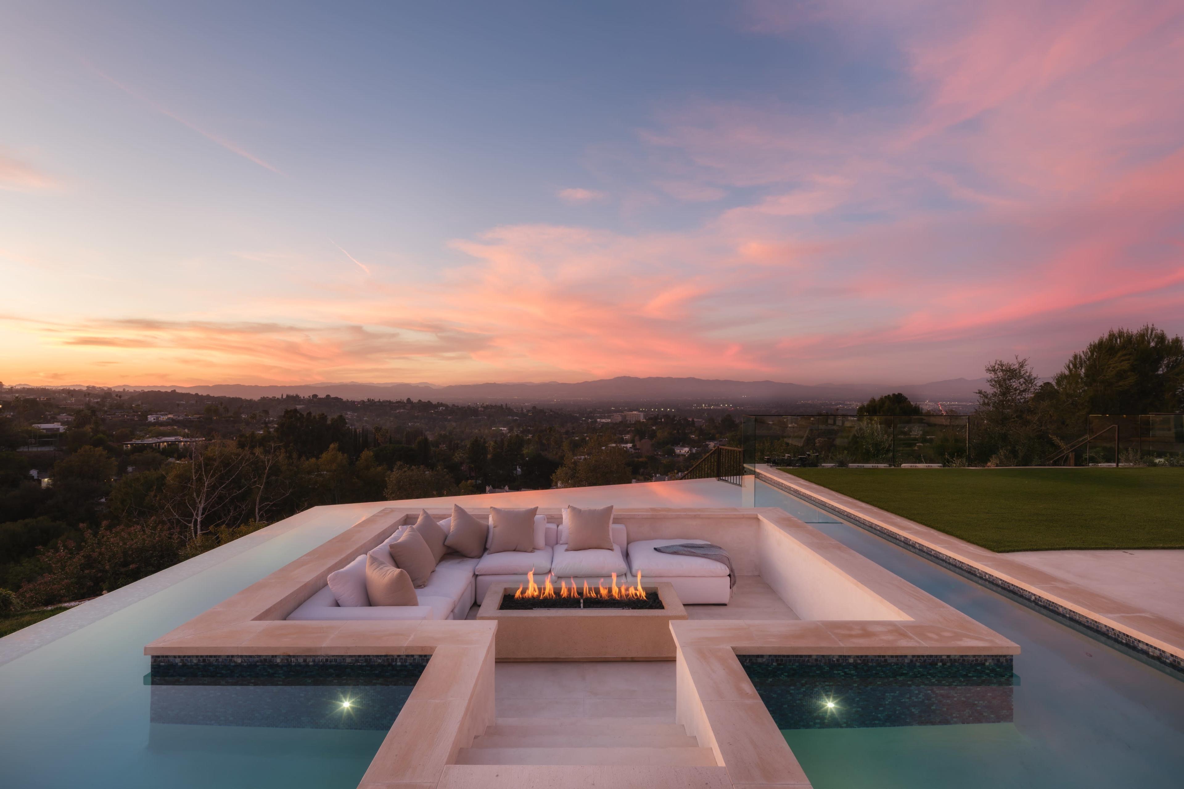 The image shows a modern outdoor seating area with a fire pit, surrounded by a swimming pool, overlooking a scenic landscape during sunset.
