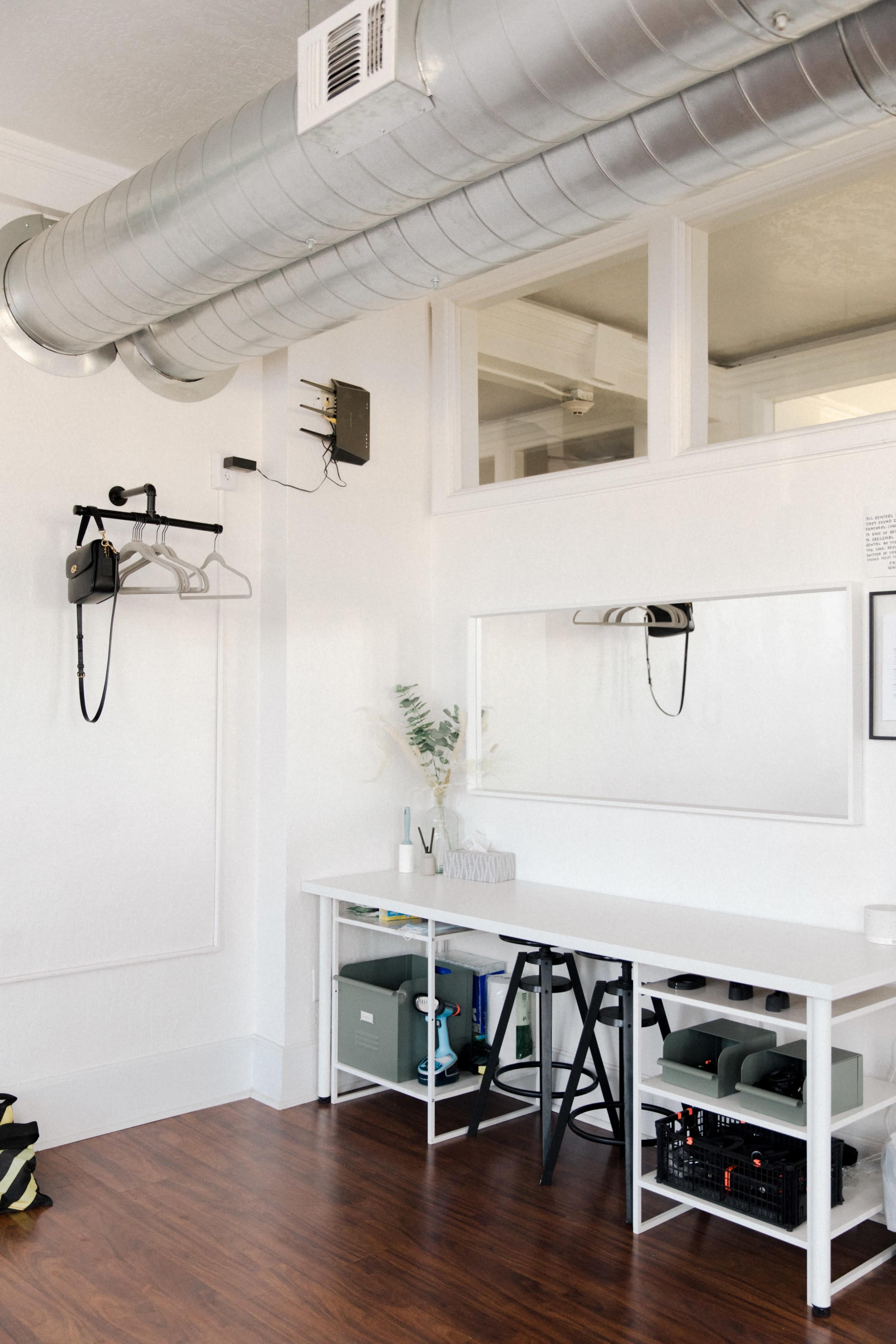 A minimalist room with a white wall, a long table with a black stool, a large mirror, and a wall-mounted hairdryer, all beneath an exposed duct.
