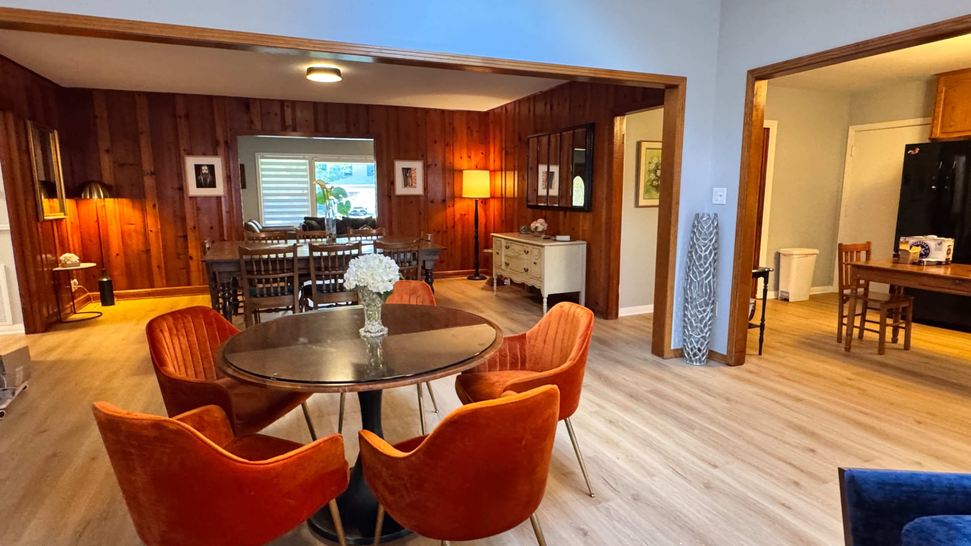 The image shows a cozy dining area with a round table surrounded by orange chairs, leading to a wooden-walled living space and an adjoining kitchen area.