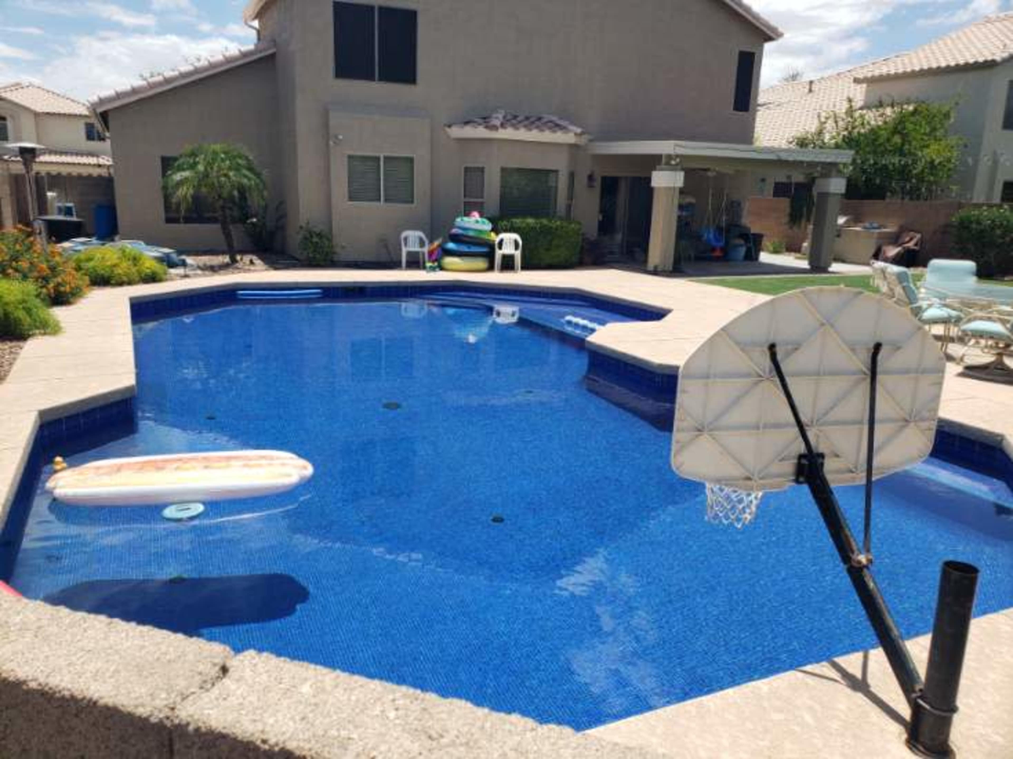 A backyard pool surrounded by patio furniture and colorful inflatable rafts under a clear sky.