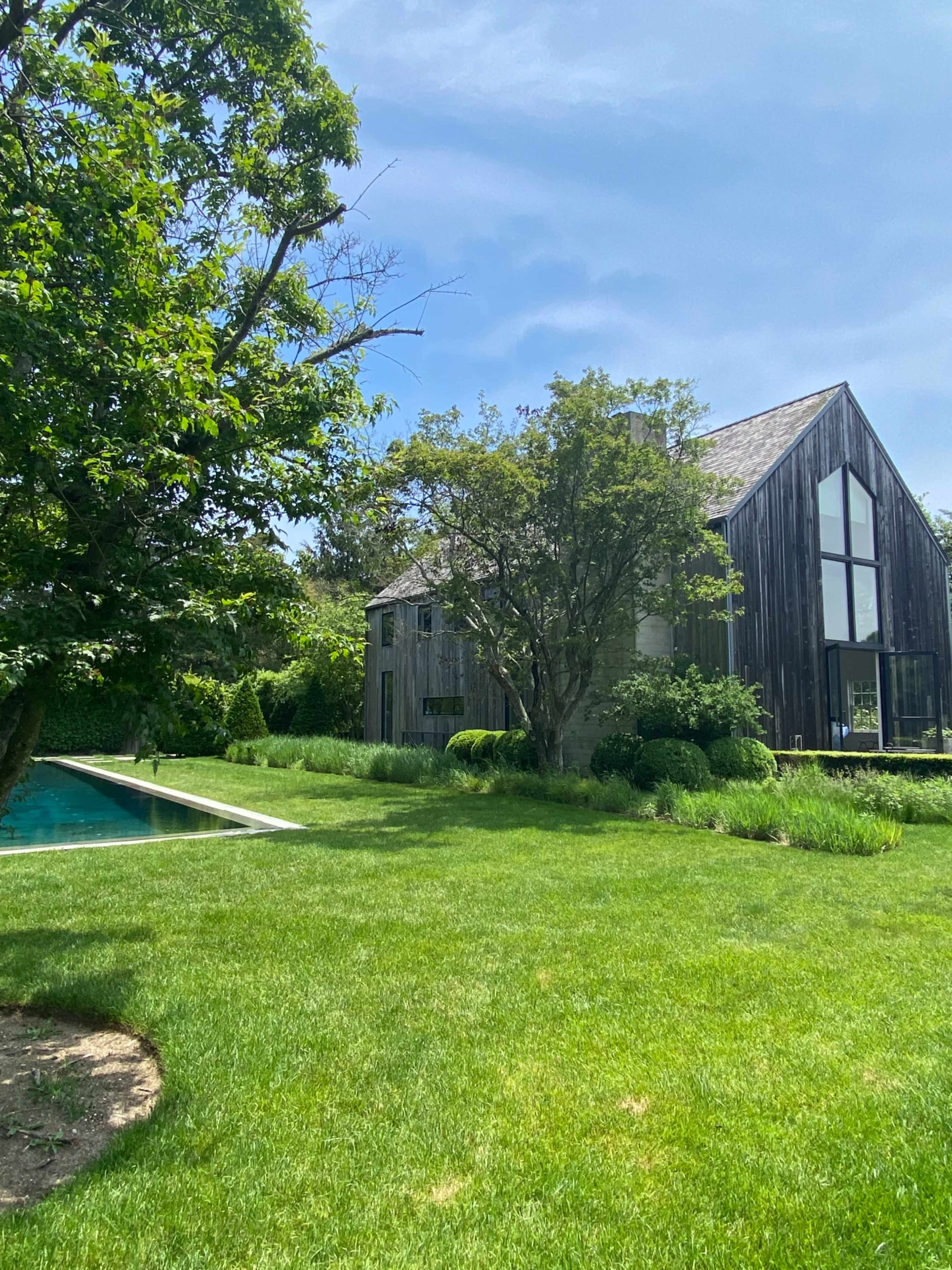 A modern wooden house with large windows stands beside a swimming pool, surrounded by lush green grass and trees.