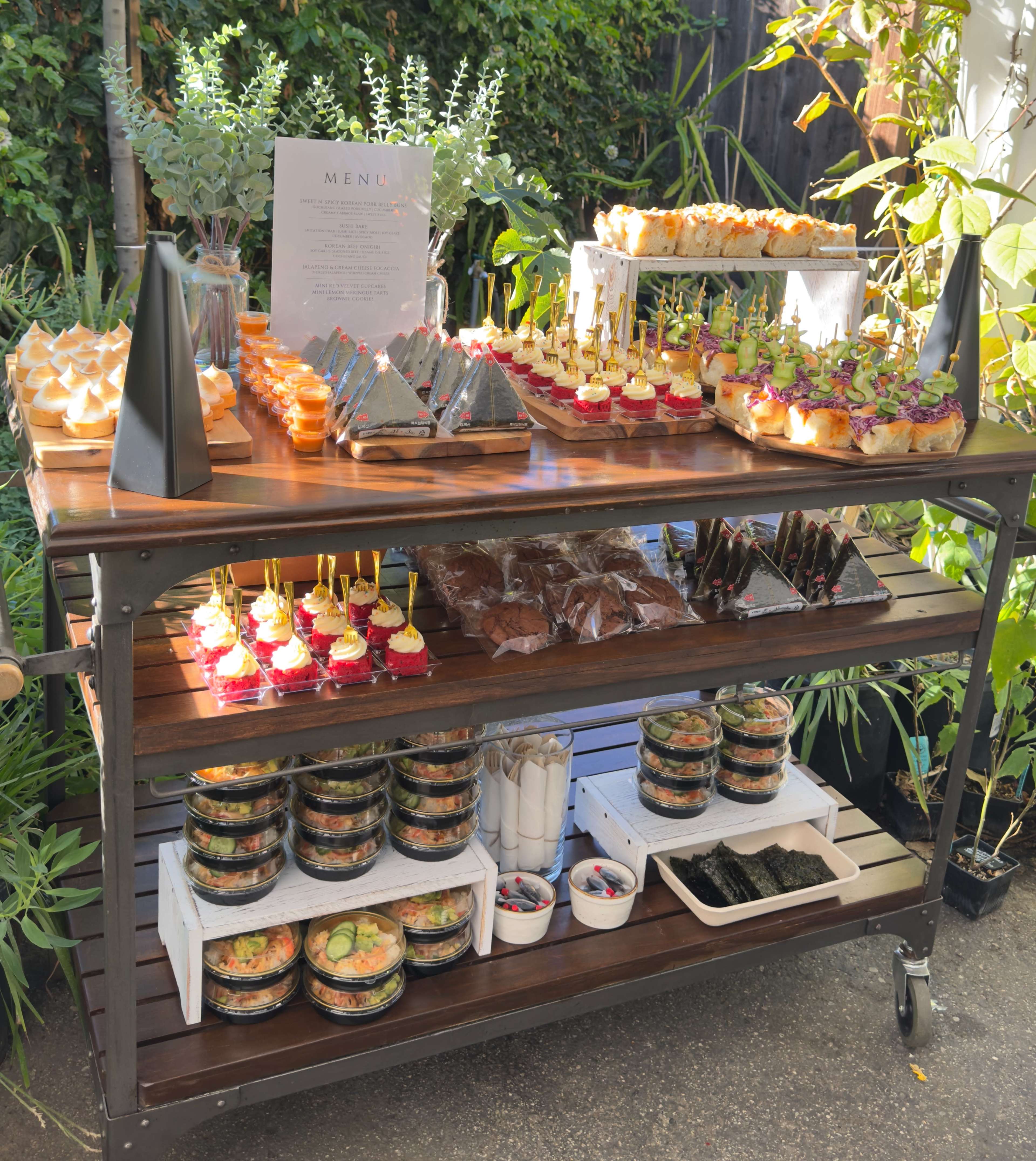 A food cart displays an assortment of neatly arranged appetizers, desserts, and salads, set against a backdrop of greenery.