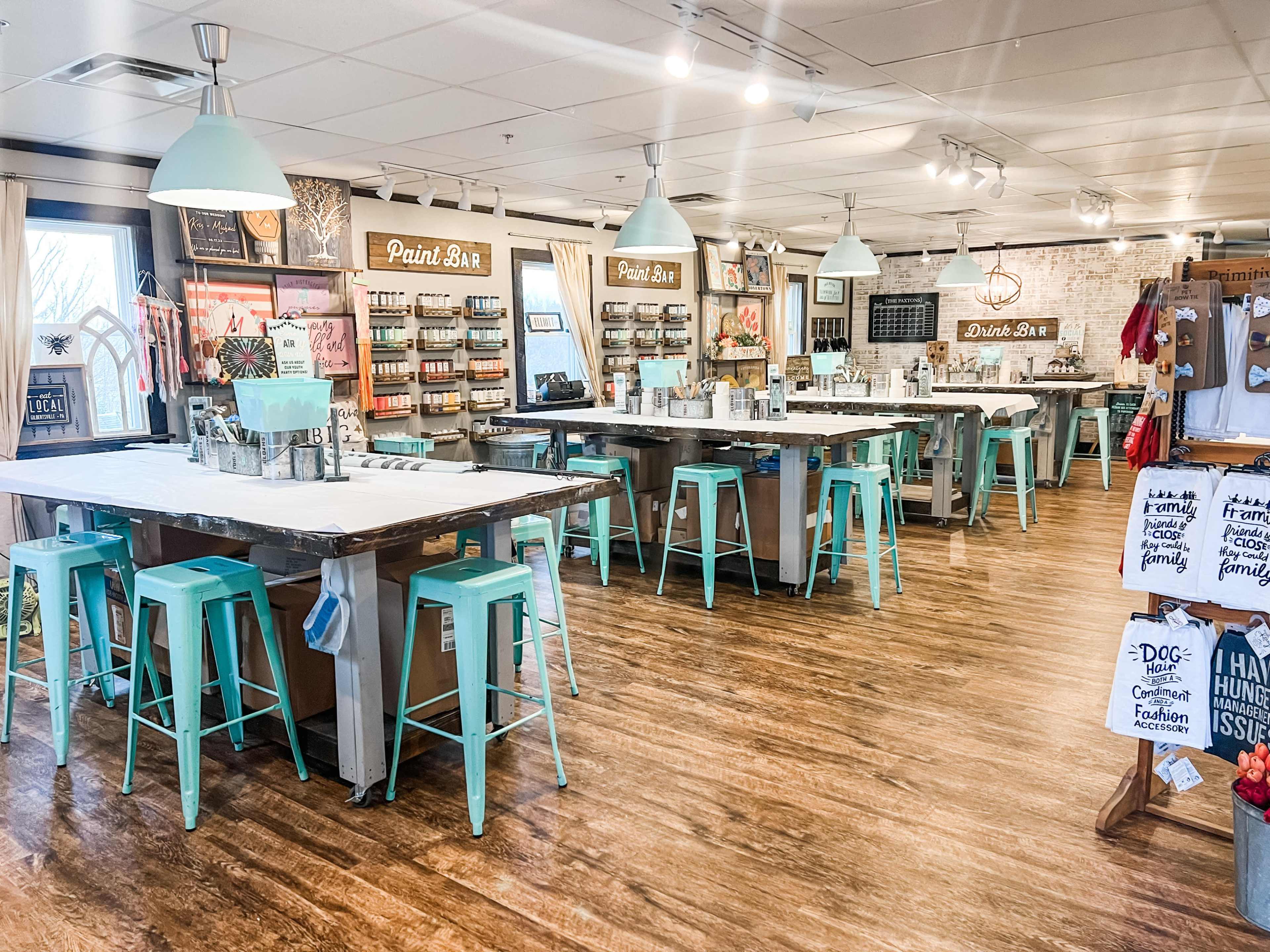 A brightly-lit craft studio featuring multiple tables with mint green stools, a paint bar, and shelves stocked with various paint supplies.